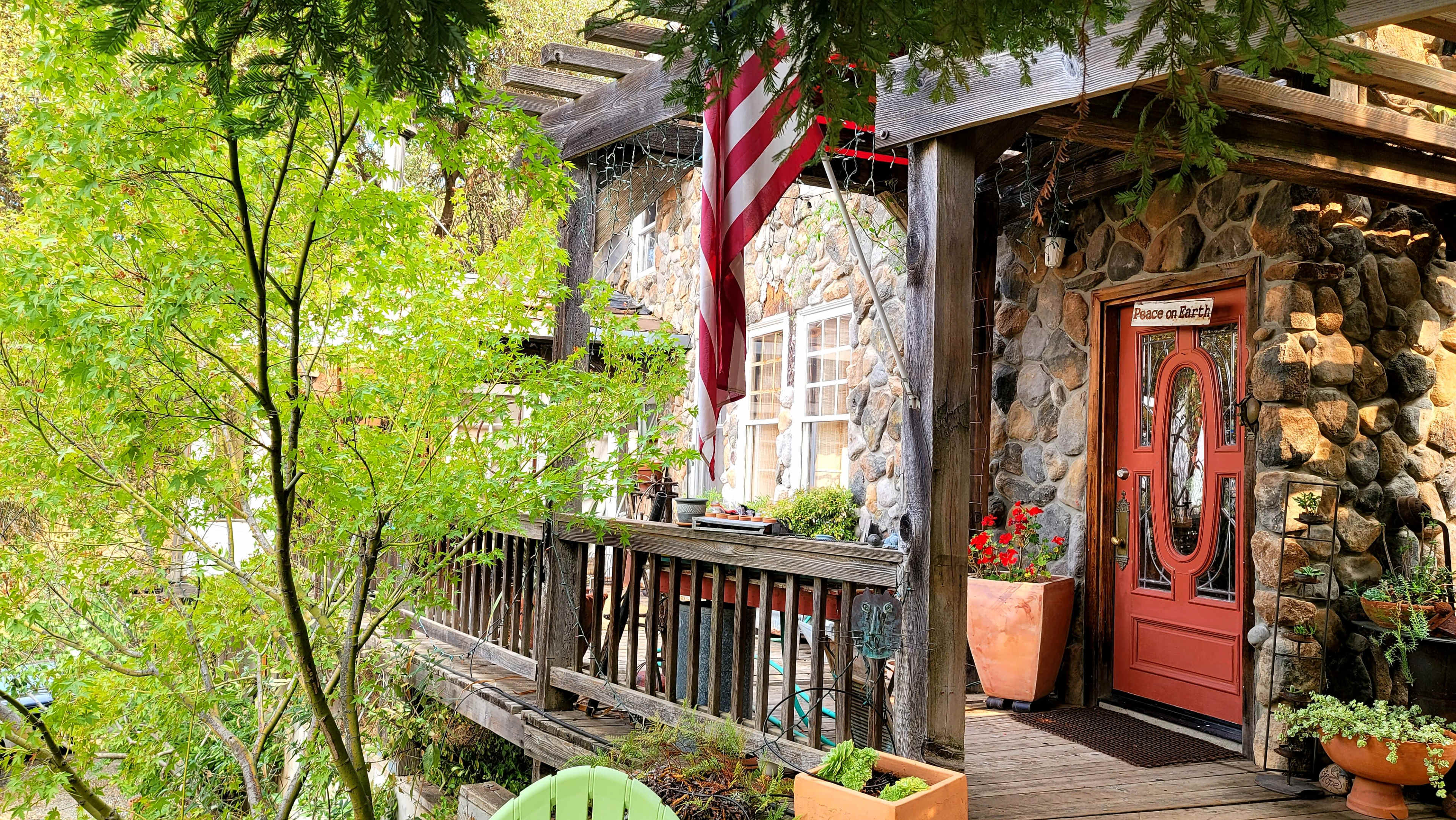 A stone cottage with a wooden deck, an American flag hanging, and potted plants on either side of the entrance.