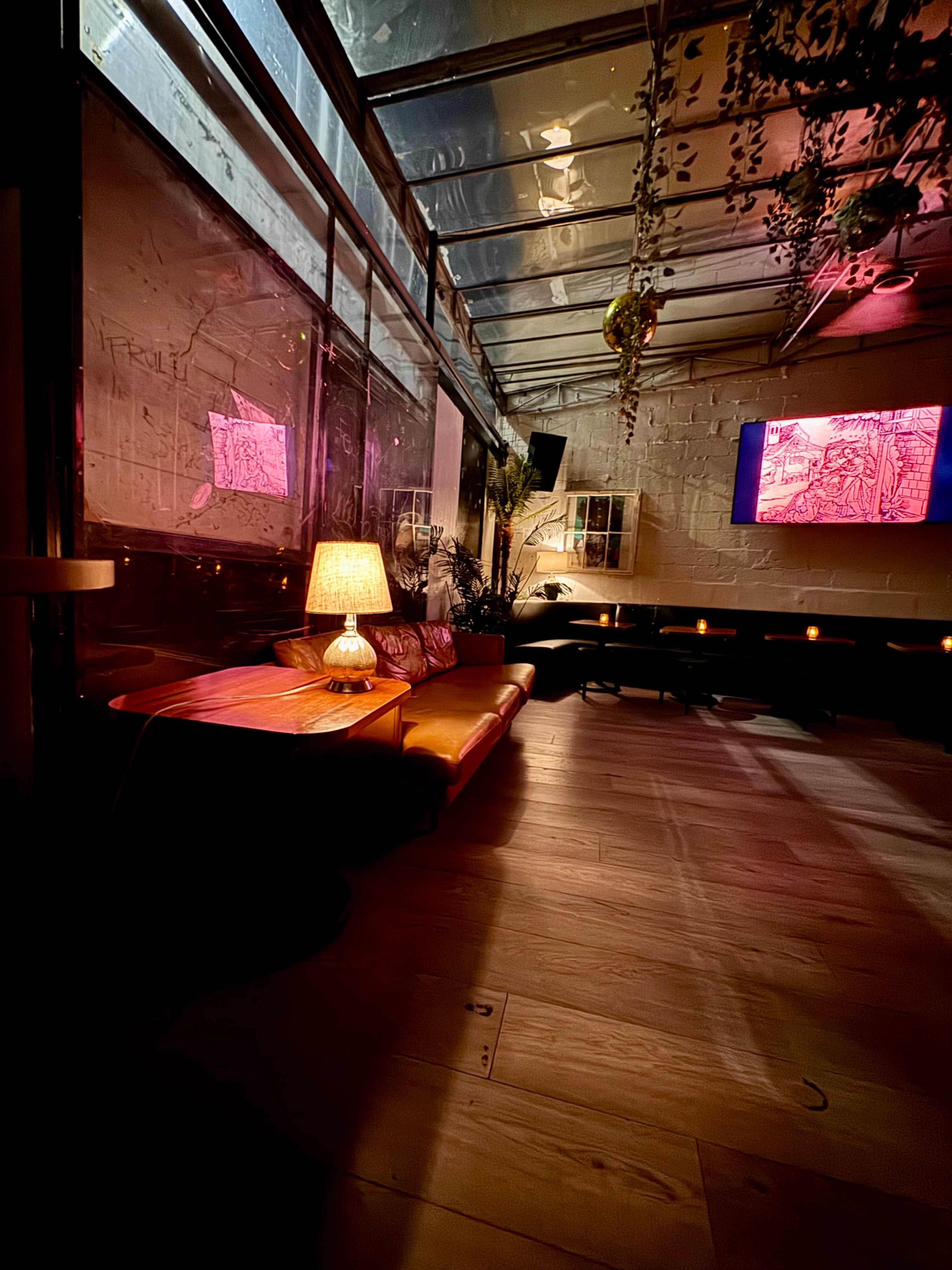 The image shows a cozy interior space with a glass ceiling, featuring a well-lit seating area, a lamp on a wooden table, and a wall-mounted screen displaying vibrant artwork.