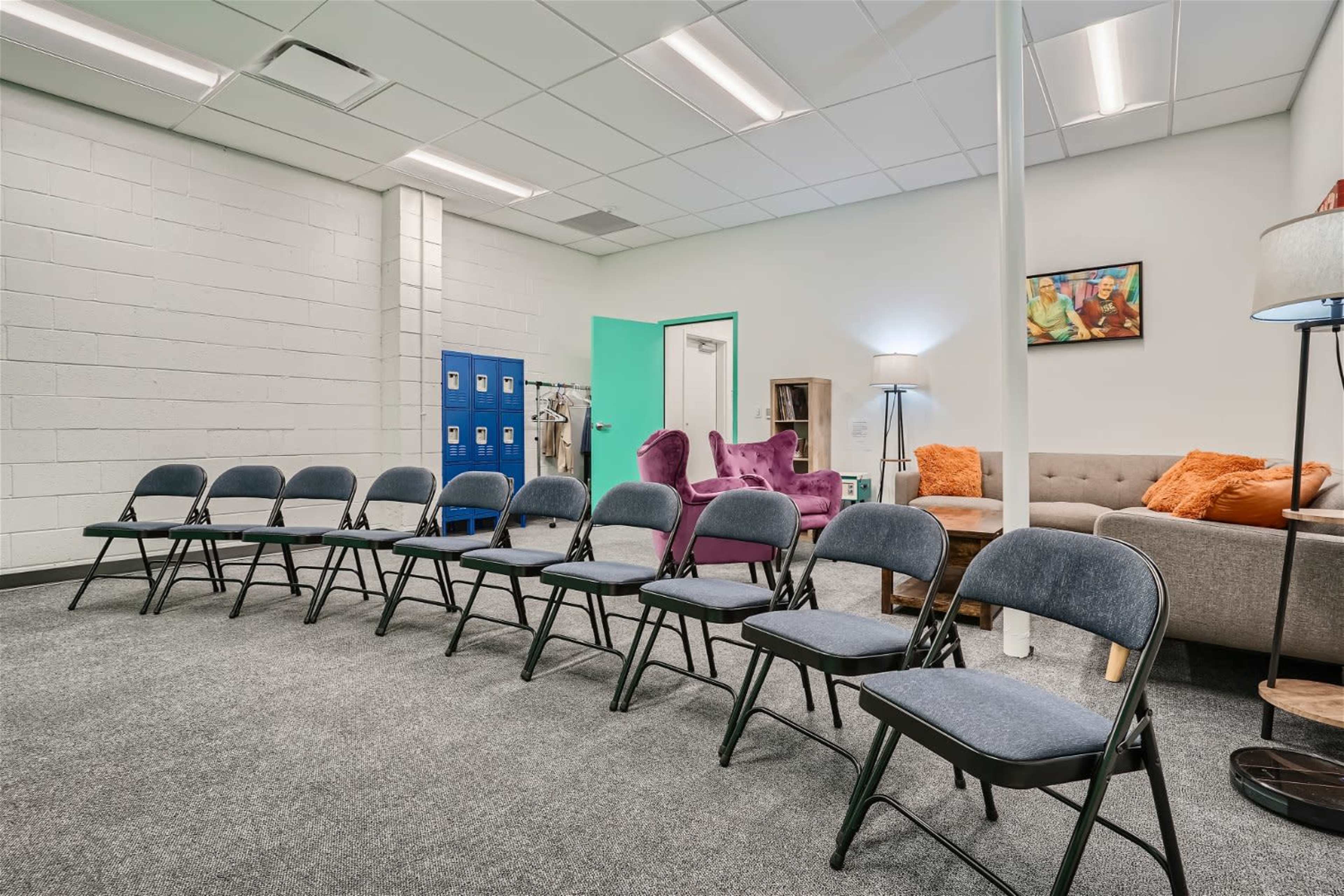 A waiting area is furnished with several folding chairs arranged in a row, a couch with orange cushions, and a blue locker against the wall.