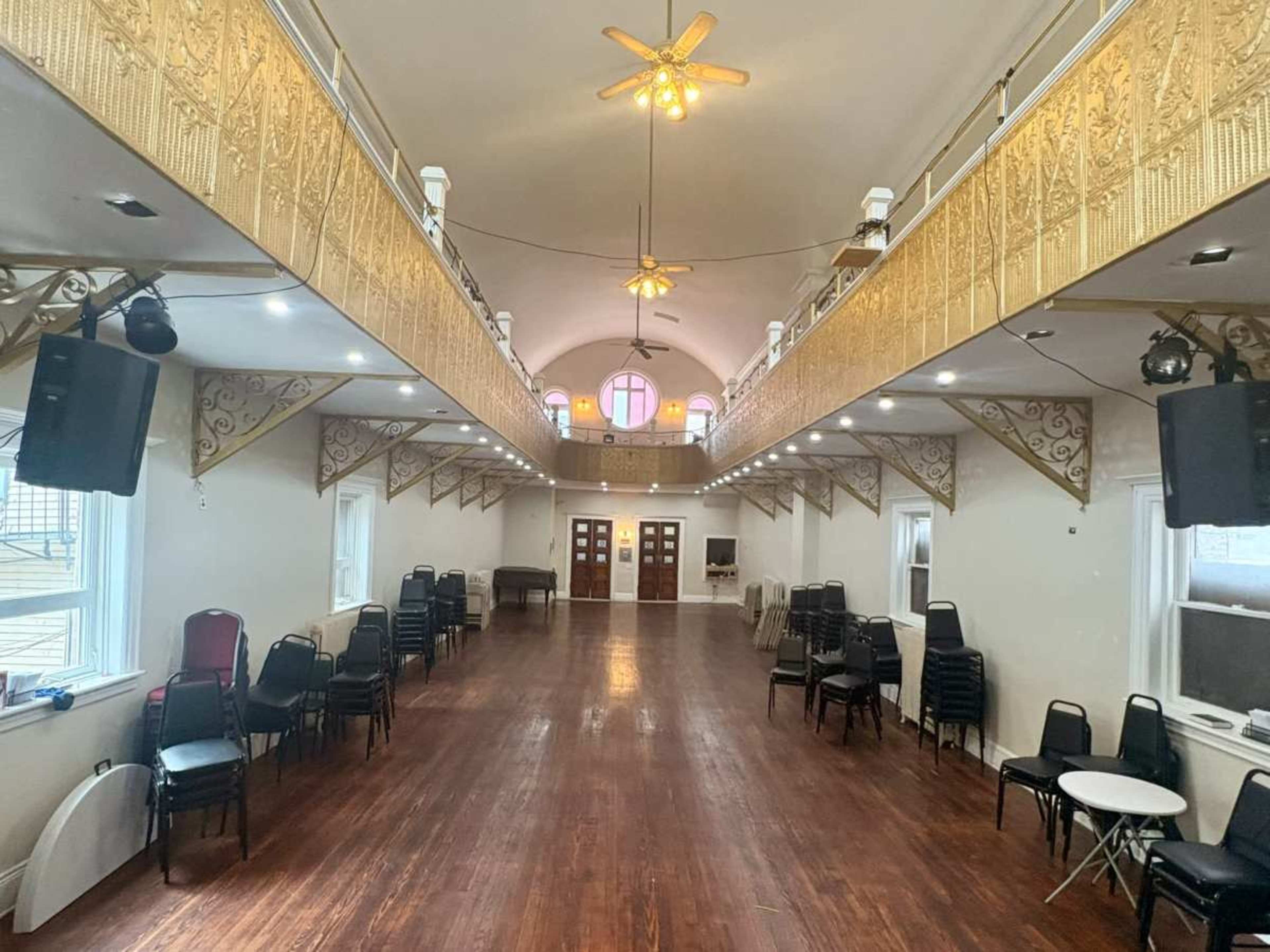 The image shows a spacious event hall with wooden flooring, two rows of balcony seating, and several tables and chairs arranged throughout the area.