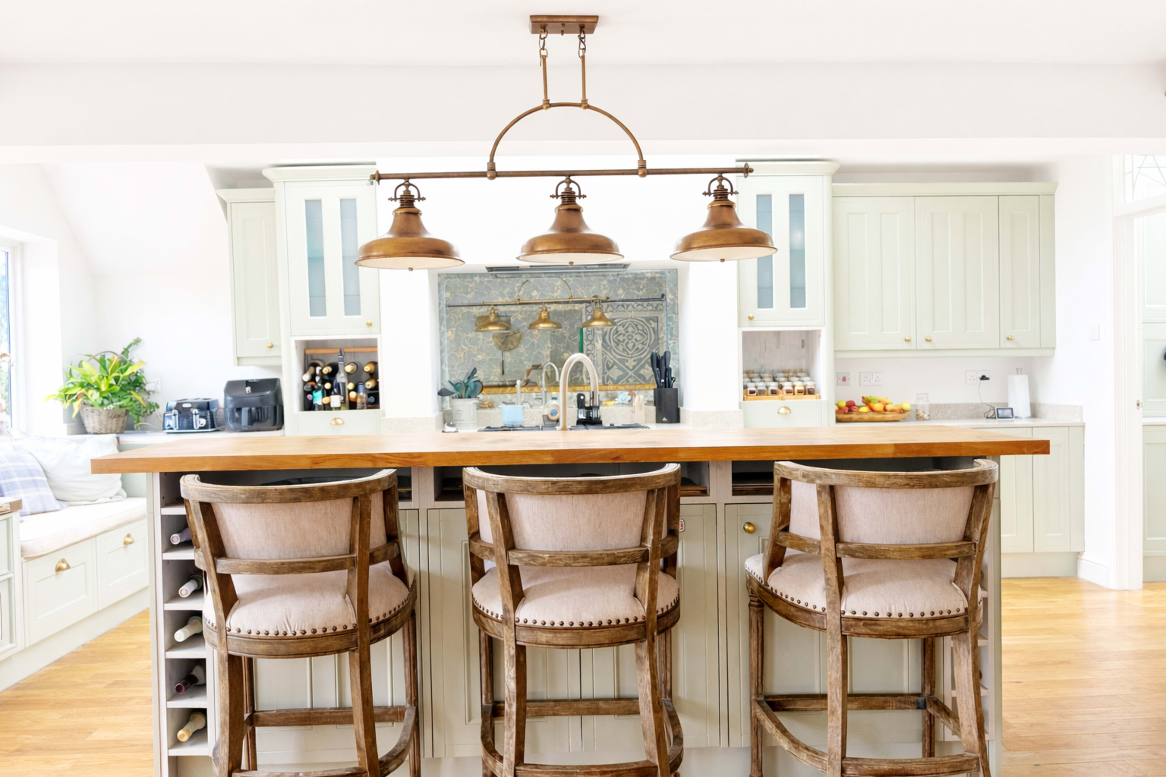 The image shows a modern kitchen featuring a central island with three wooden bar stools, pendant lighting above, and cabinetry with a light green hue.