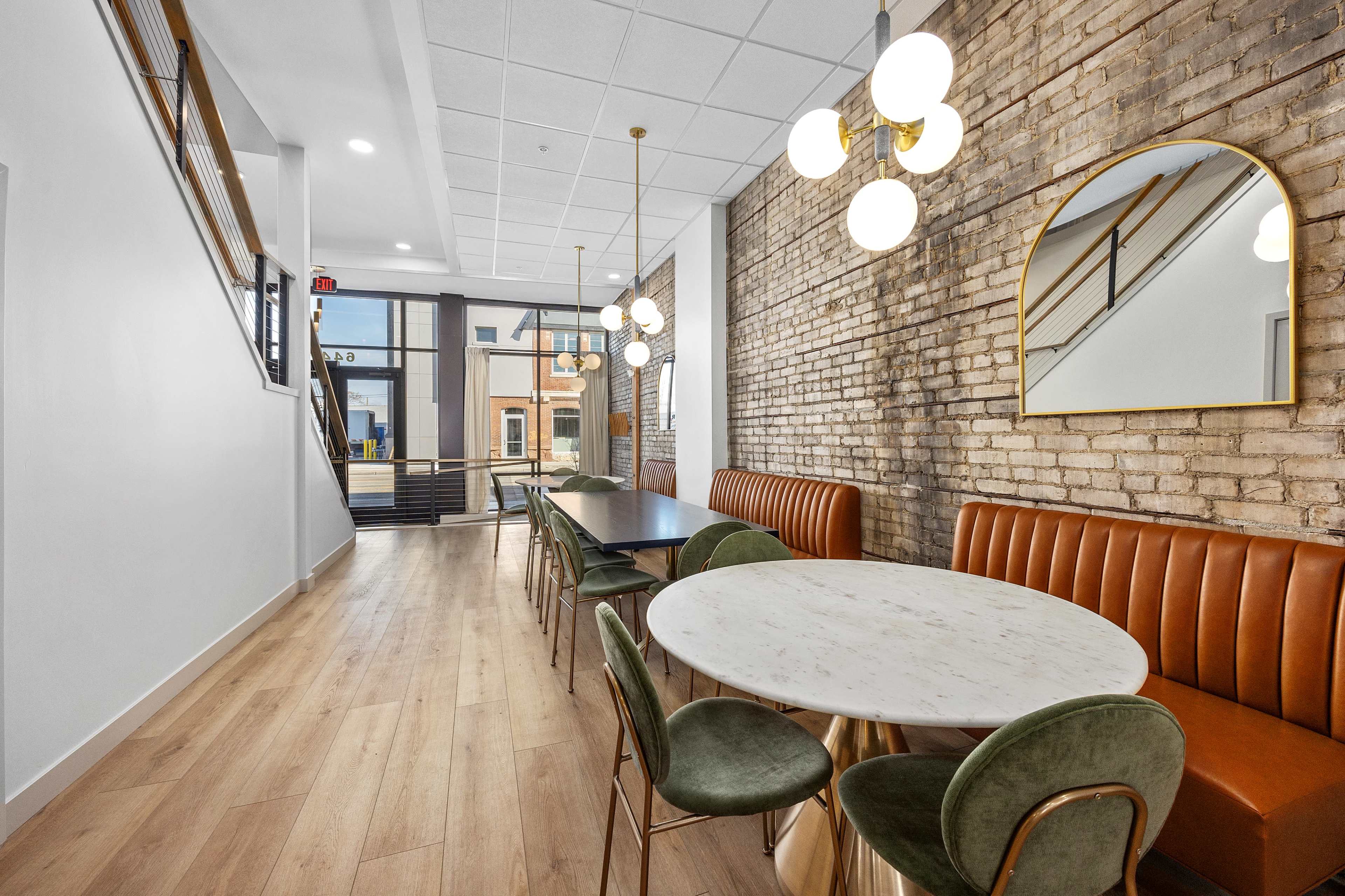 A modern dining area featuring a round marble table, green velvet chairs, a brown leather banquette, and a staircase in a space with exposed brick walls.