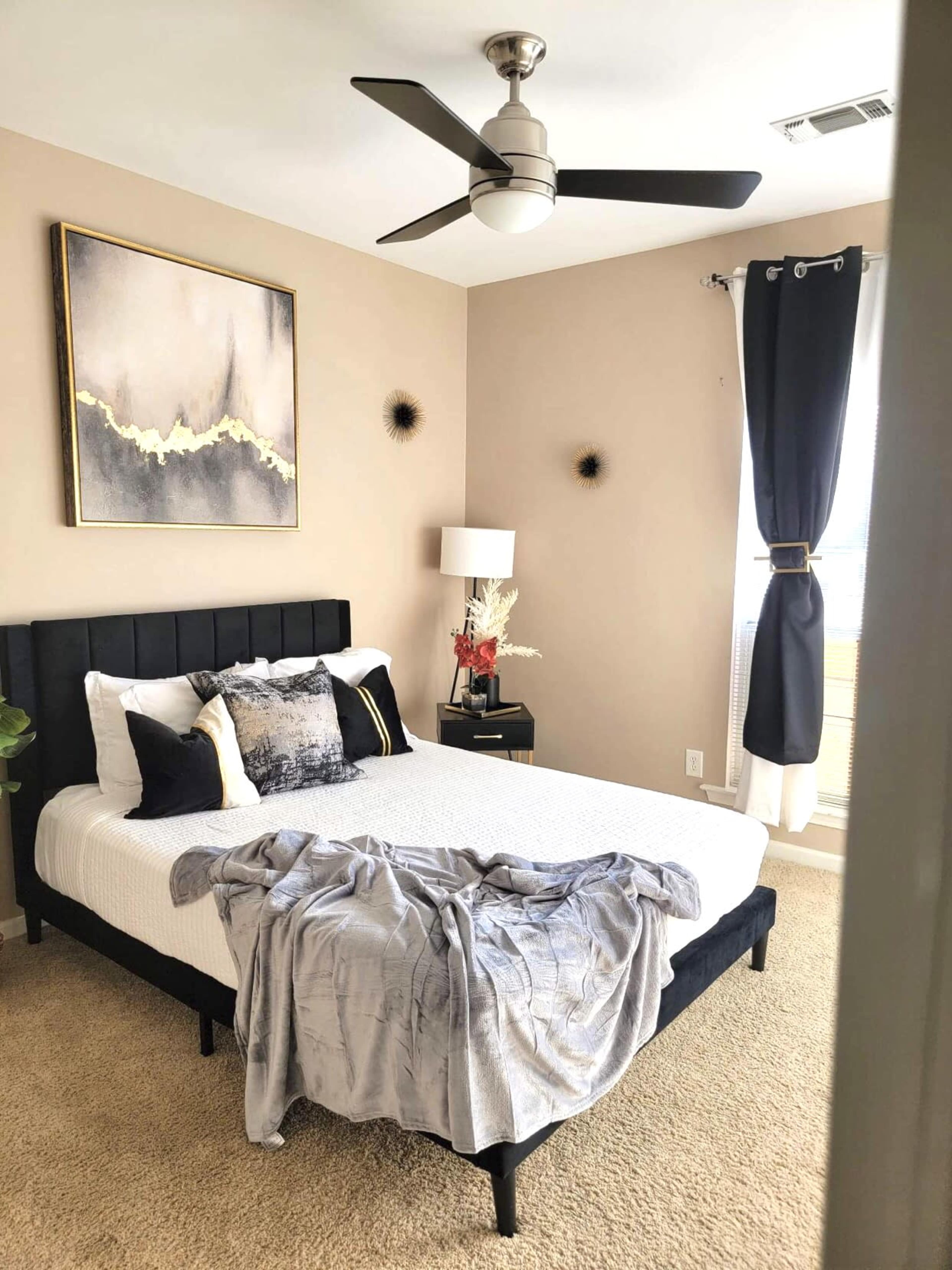 A neatly arranged bedroom features a bed with a light gray throw, a bedside table with a lamp, and modern art on the wall.