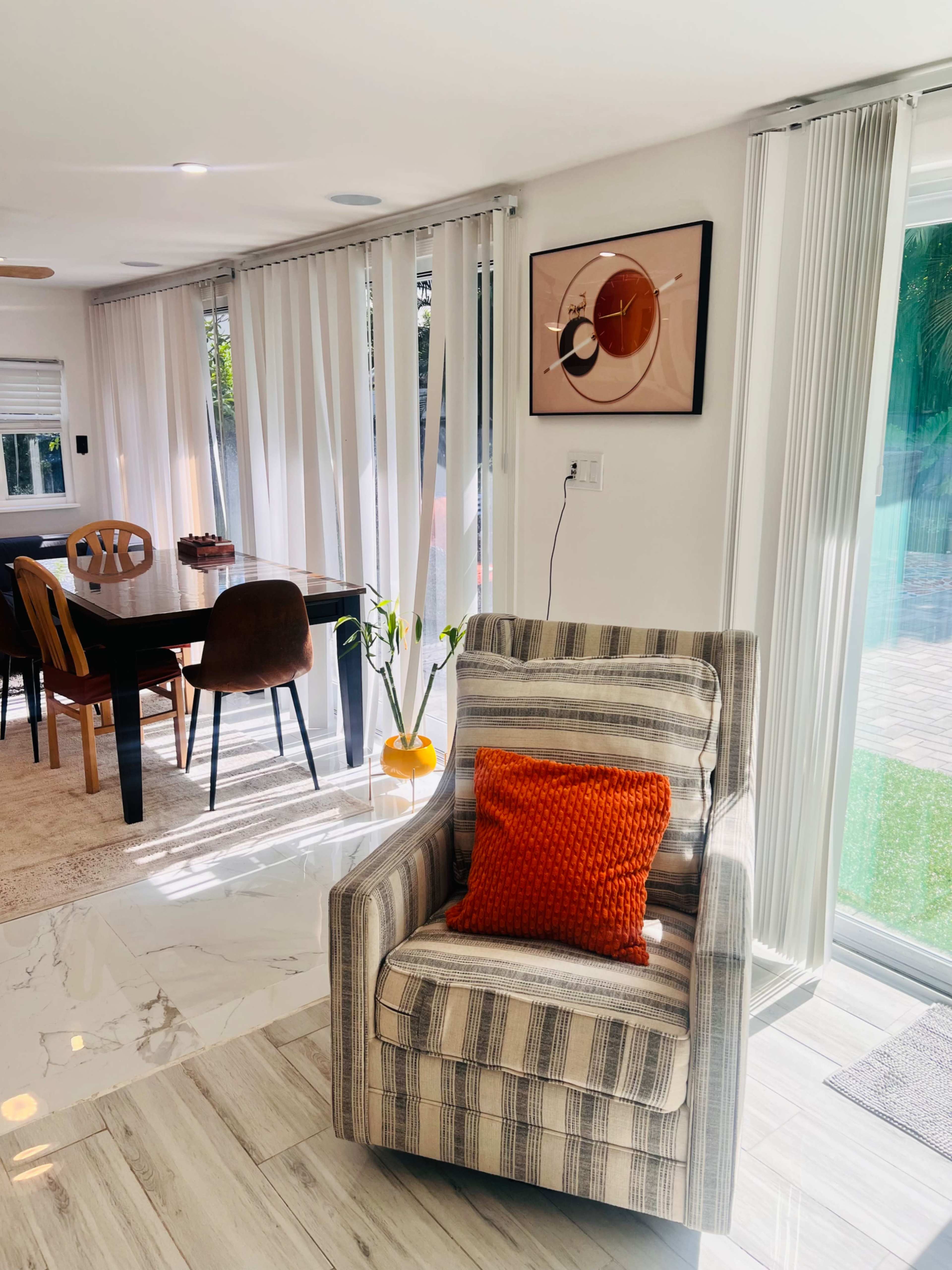 A striped armchair with an orange pillow is positioned near a table and chairs in a bright, airy living space featuring large windows and sliding doors.