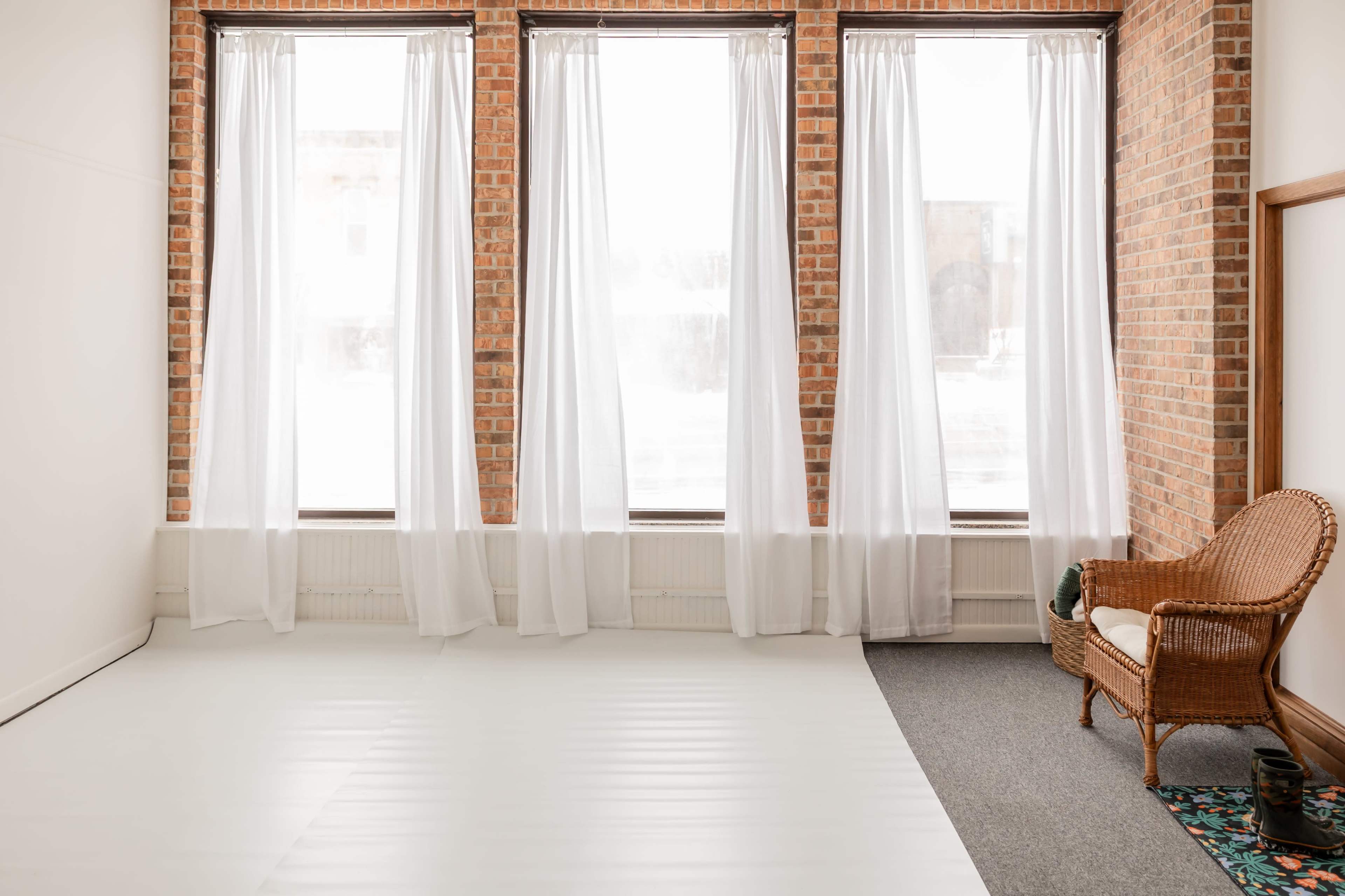 The image shows a spacious room with three large windows covered by sheer white curtains, featuring a light-colored floor and a wicker chair in one corner.