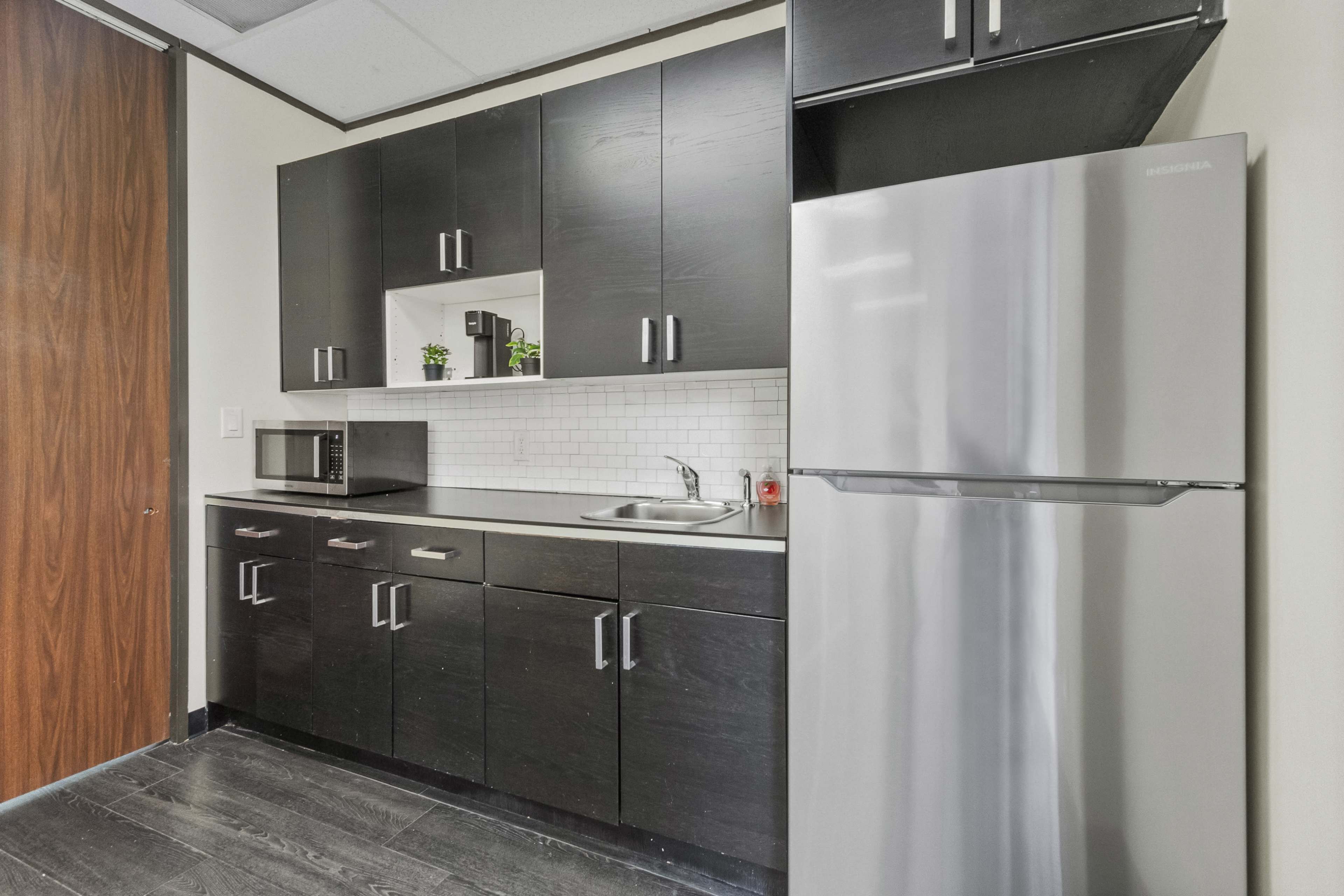 The image shows a modern kitchenette with black cabinets, a stainless steel refrigerator, a sink, a microwave, and white tile backsplash.