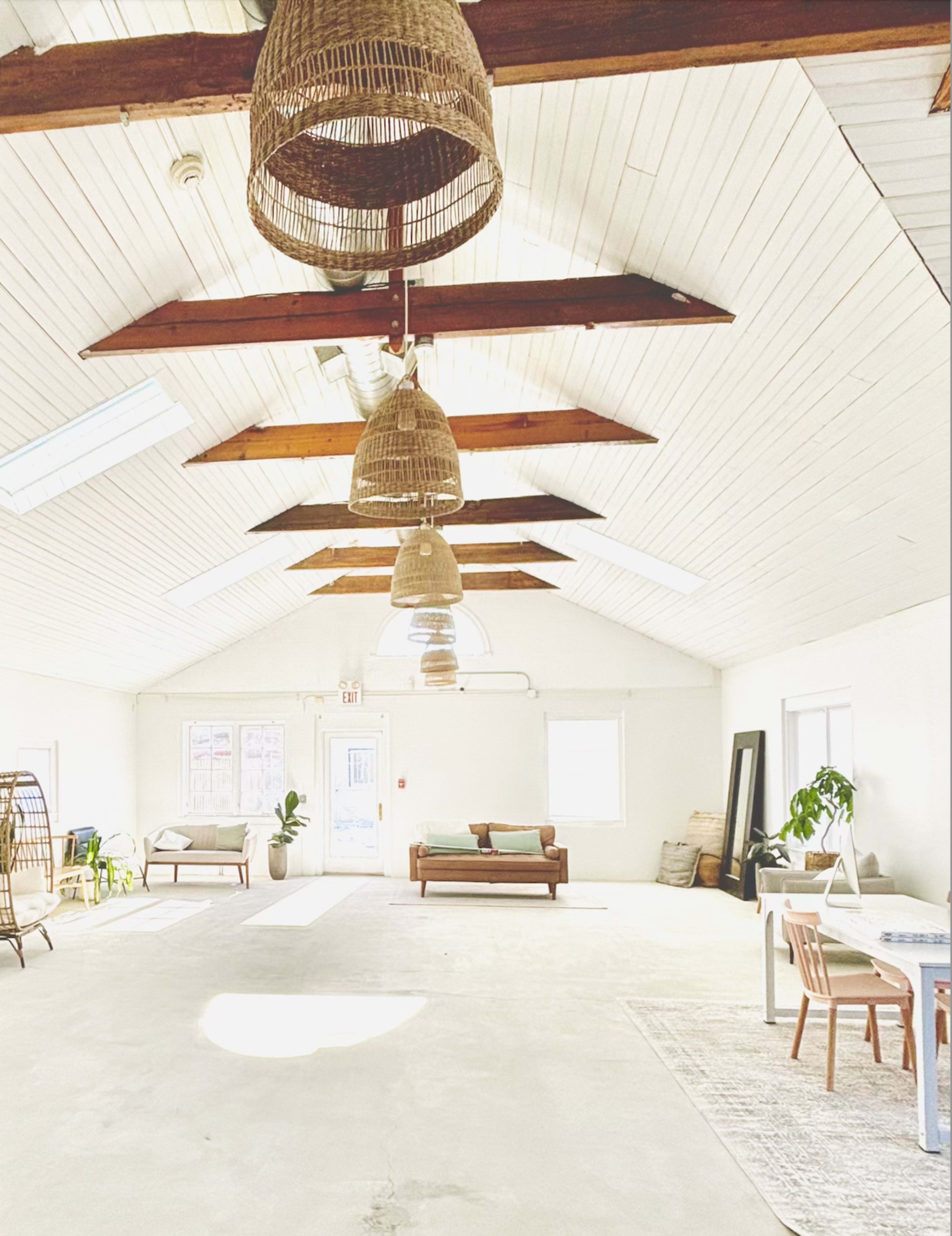 The image shows a spacious, well-lit interior with high ceilings, hanging light fixtures, and minimal furnishings, including a couch and a dining table.