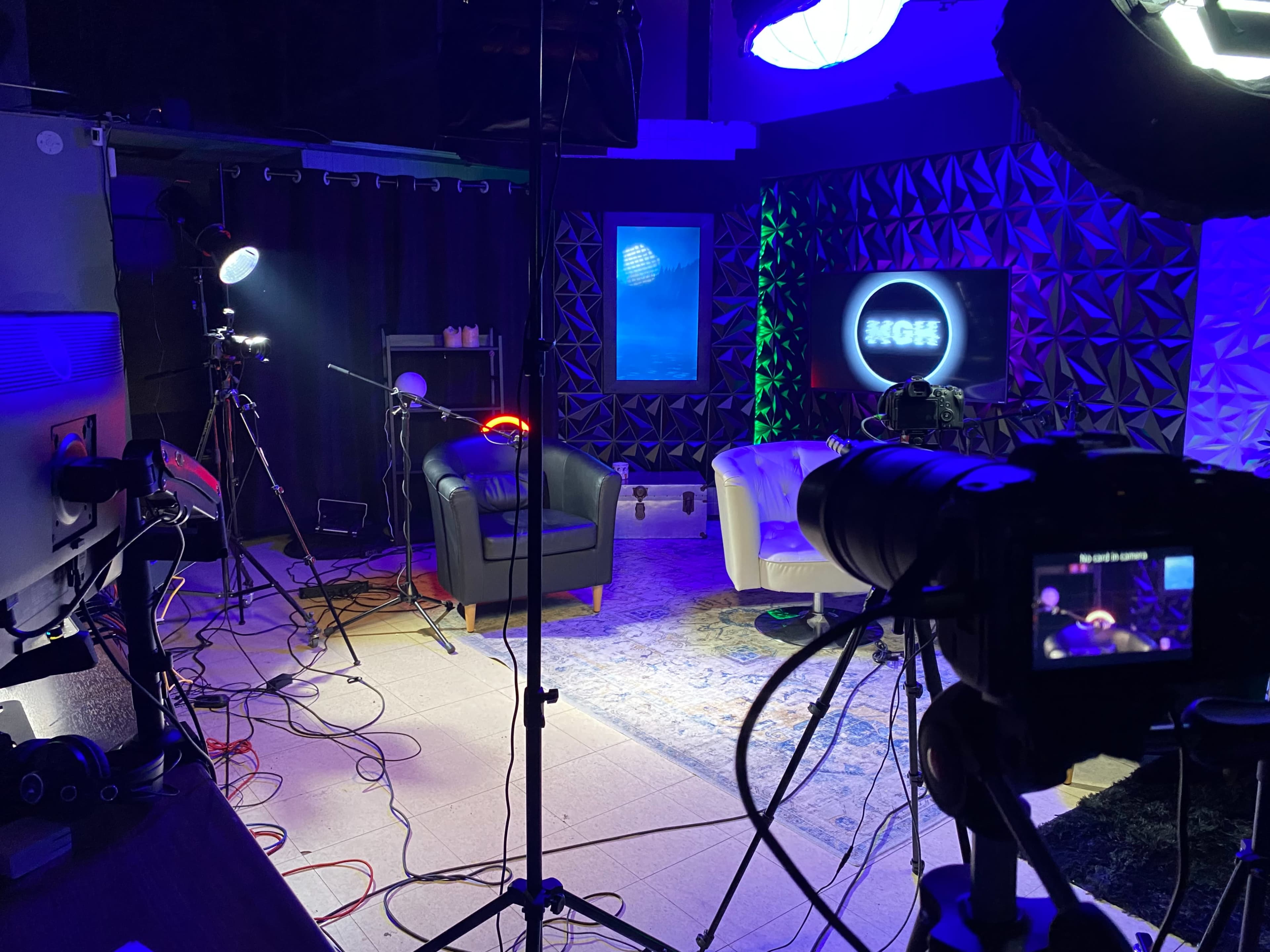 The image shows a television studio setup with two chairs, various lighting equipment, and cameras positioned for a recording.