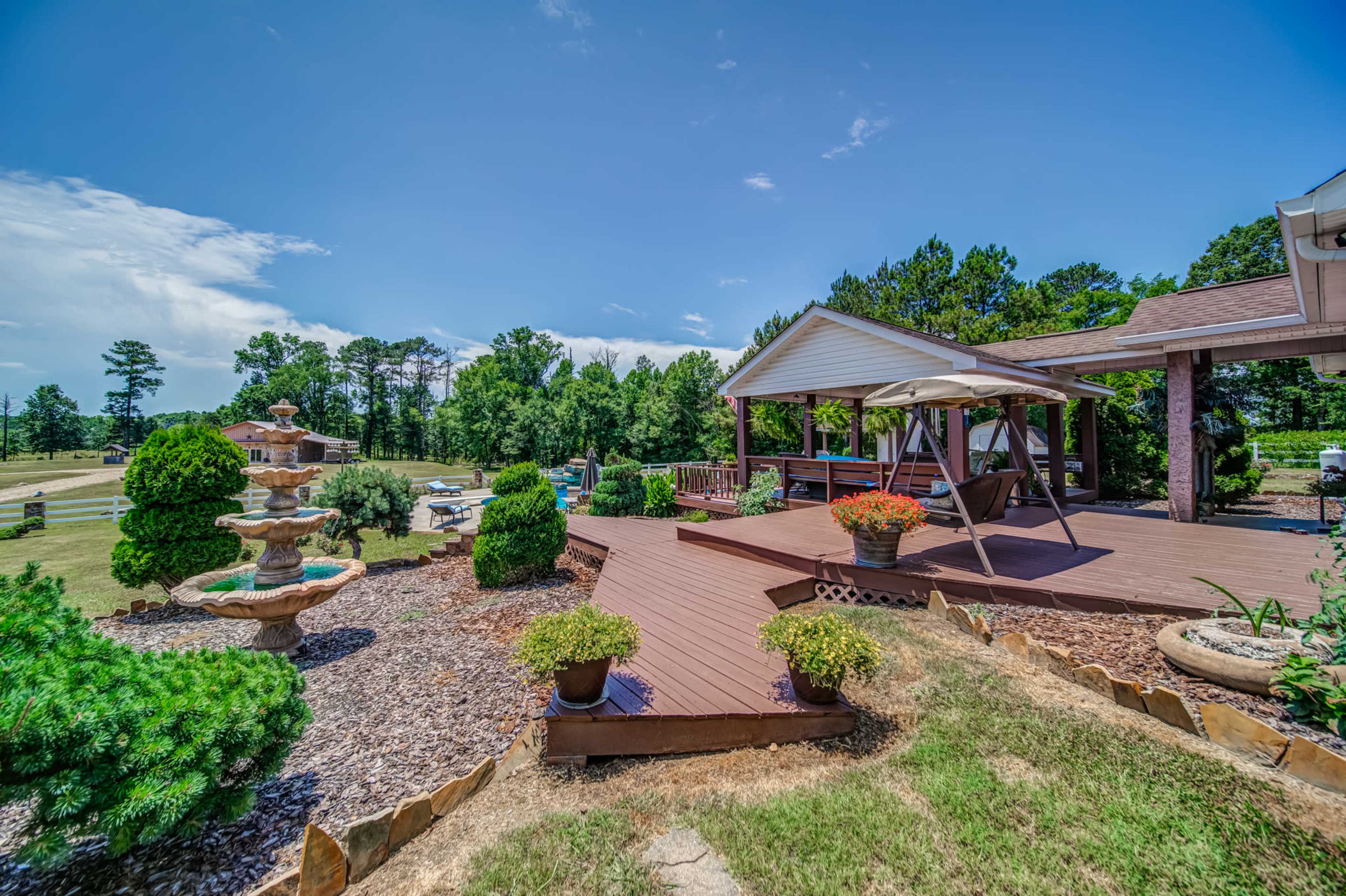 A wooden deck with potted plants and a large umbrella overlooks a landscaped yard featuring a stone fountain and a line of trees in the background.