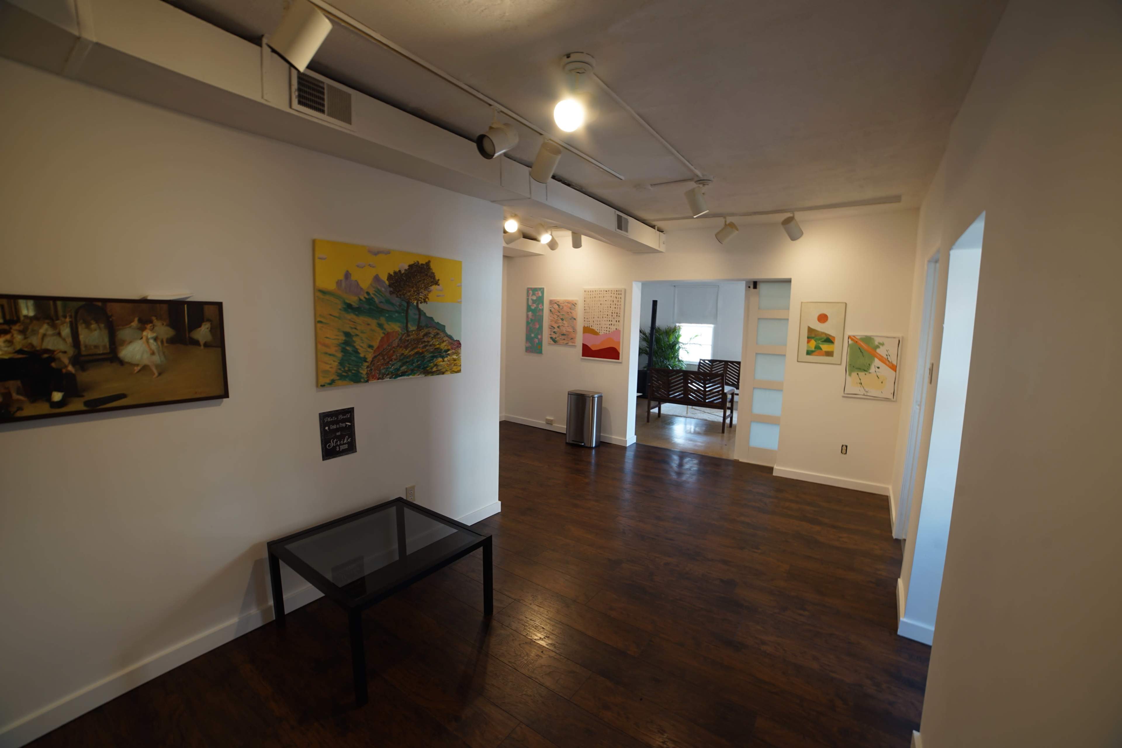 A well-lit gallery with several paintings on the white walls and a glass table in the foreground.