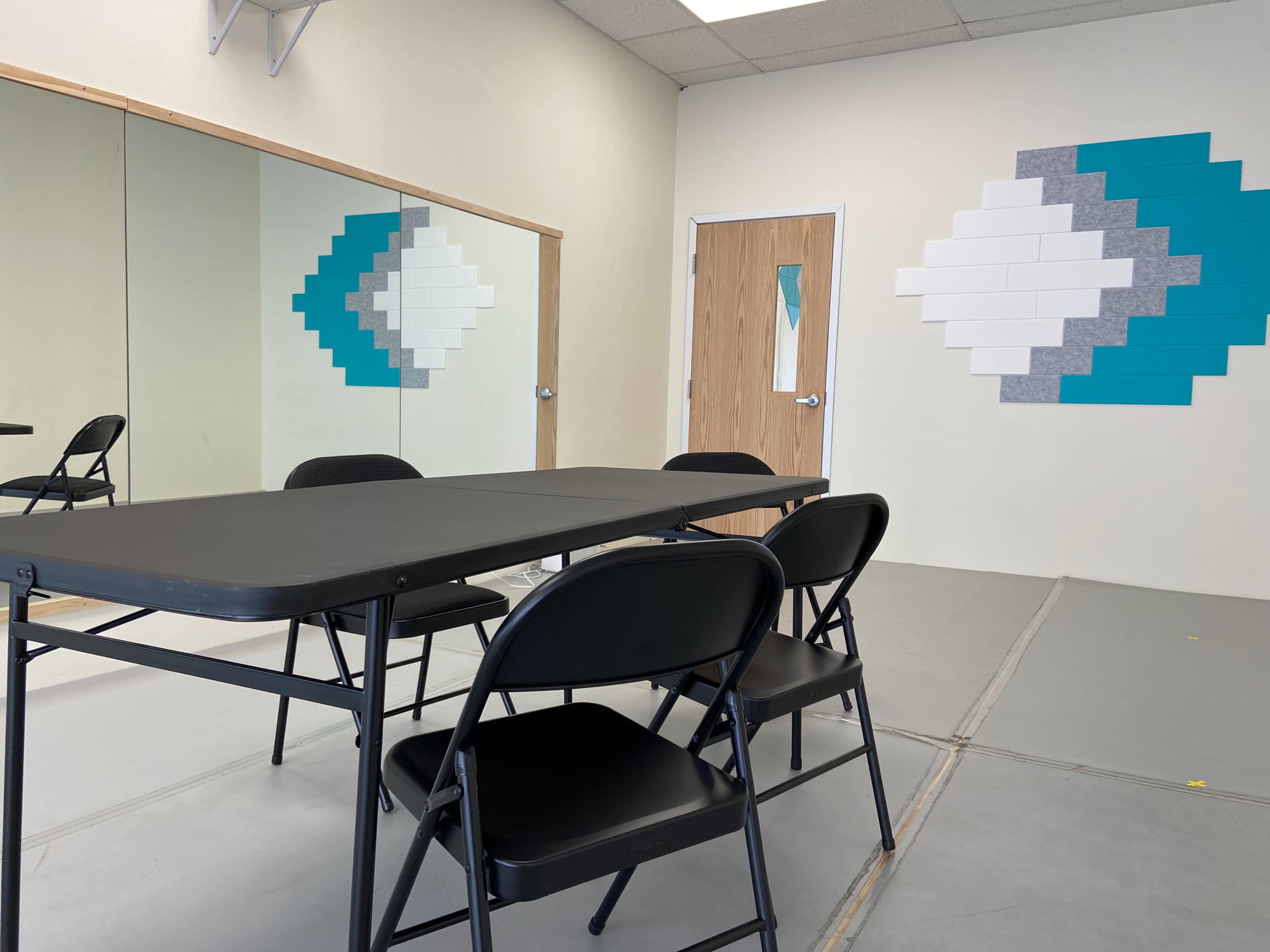 A room featuring a black folding table surrounded by four chairs, with a mirrored wall and a geometric wall decoration in teal and gray.