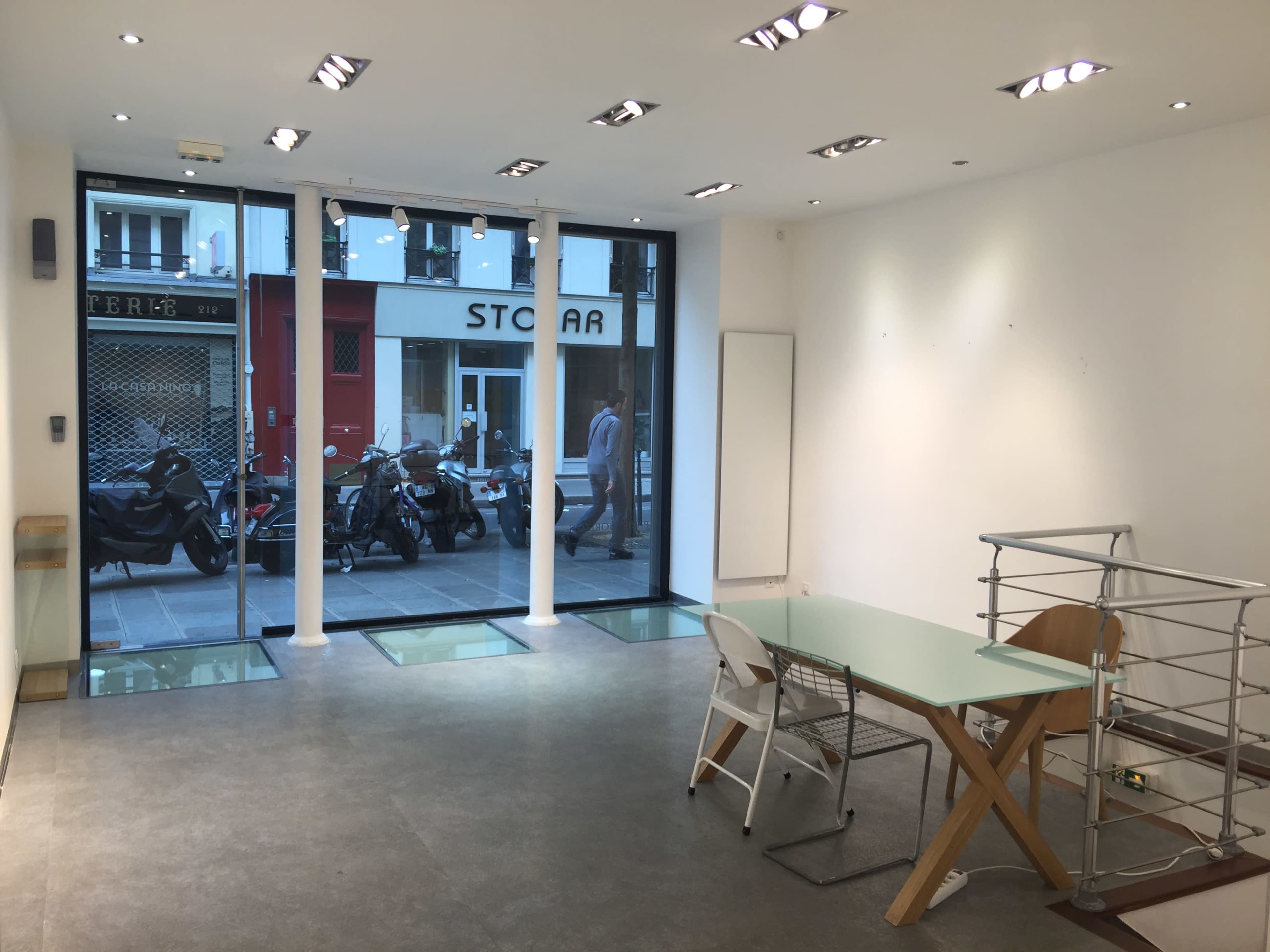 The image shows a minimalistic indoor space with large windows displaying a street scene outside, featuring parked motorcycles and pedestrians.