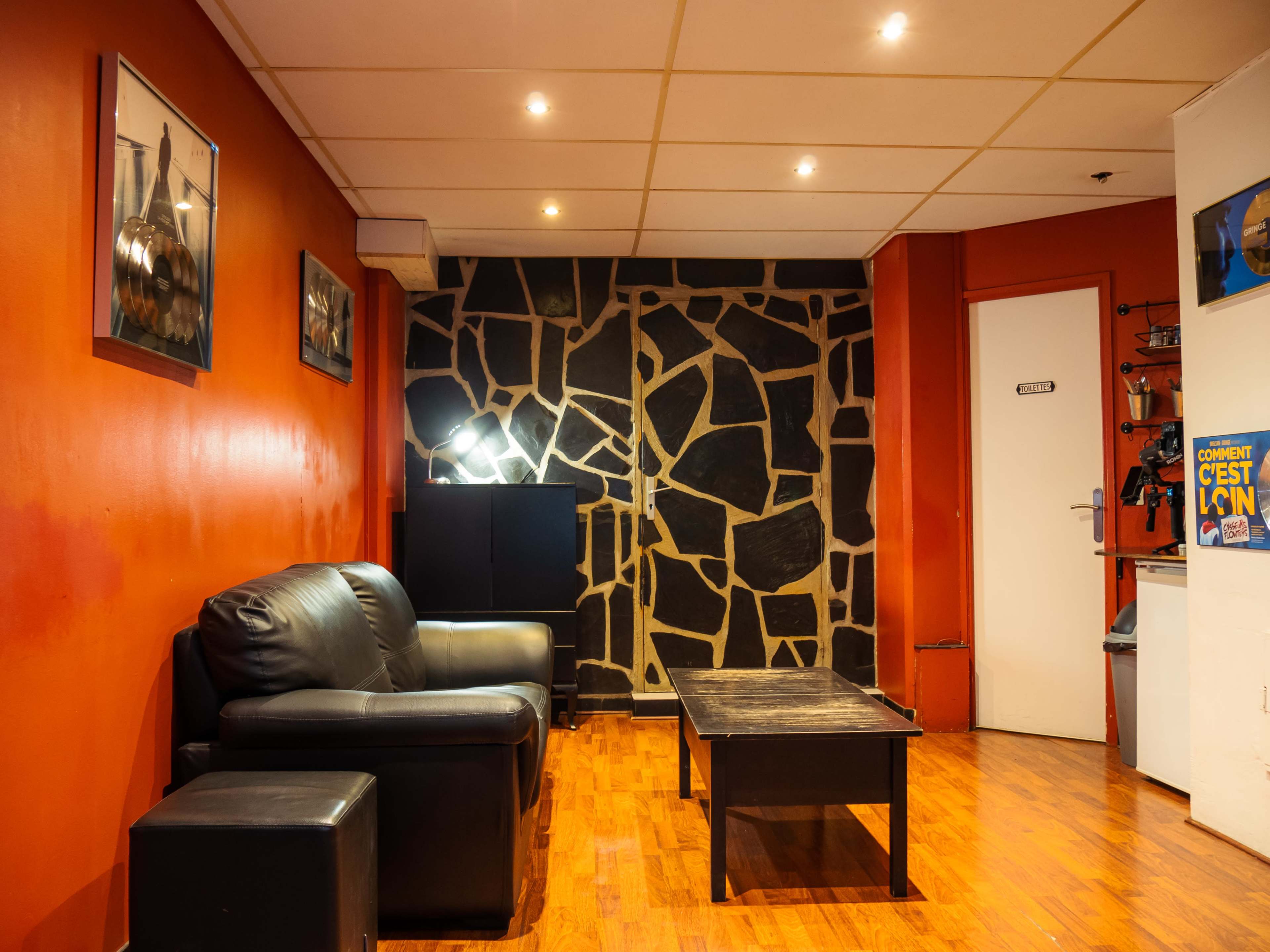 A room with an orange wall, a stone-patterned accent wall, a black leather couch, a wooden coffee table, and a door on the right.