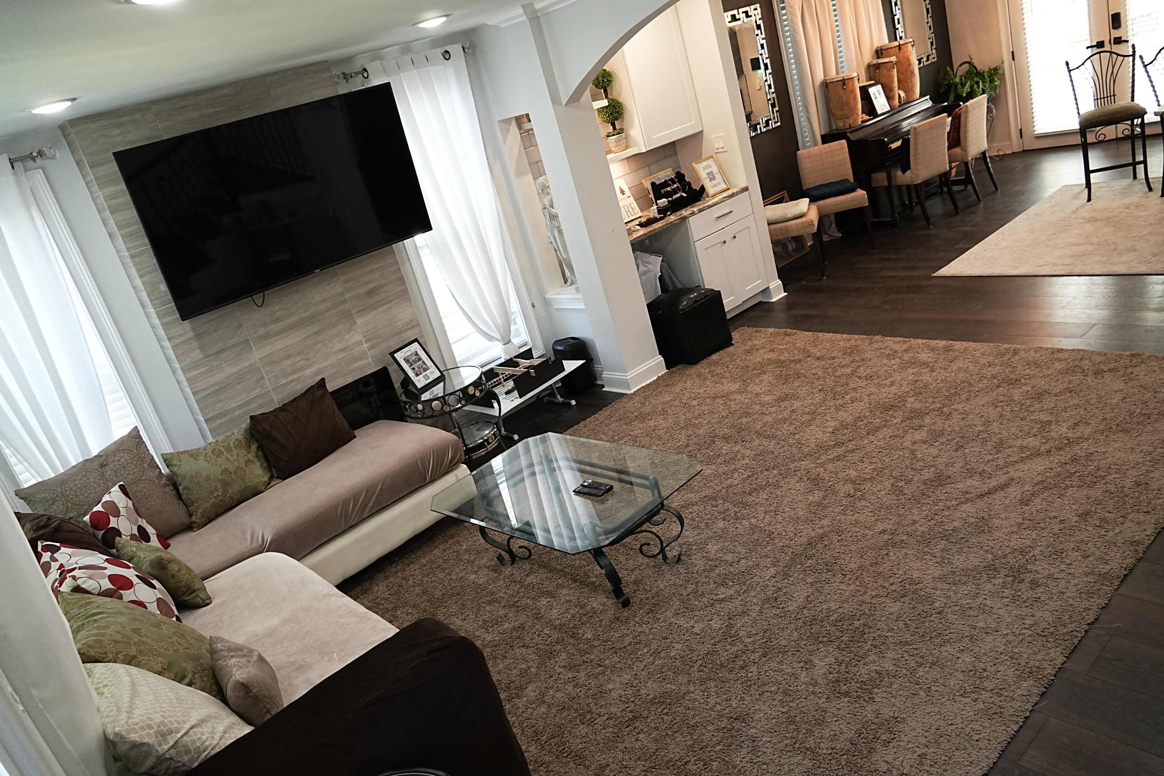 A living room features a large sectional sofa, a glass coffee table, and a television mounted on the wall, with a dining area visible in the background.