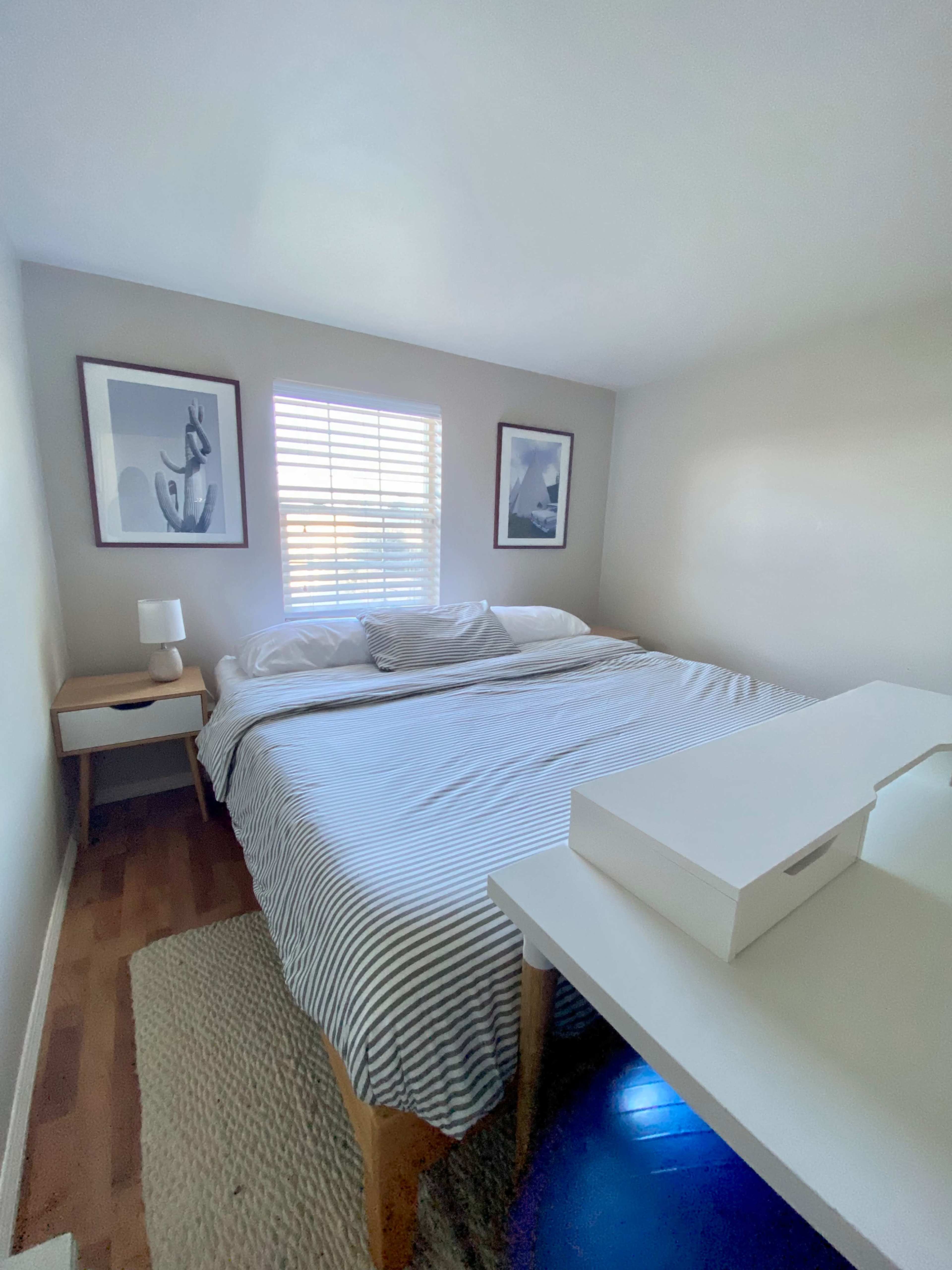 A neatly arranged bedroom features a bed with striped bedding, a bedside table with a lamp, and a white desk positioned under a window with blinds.