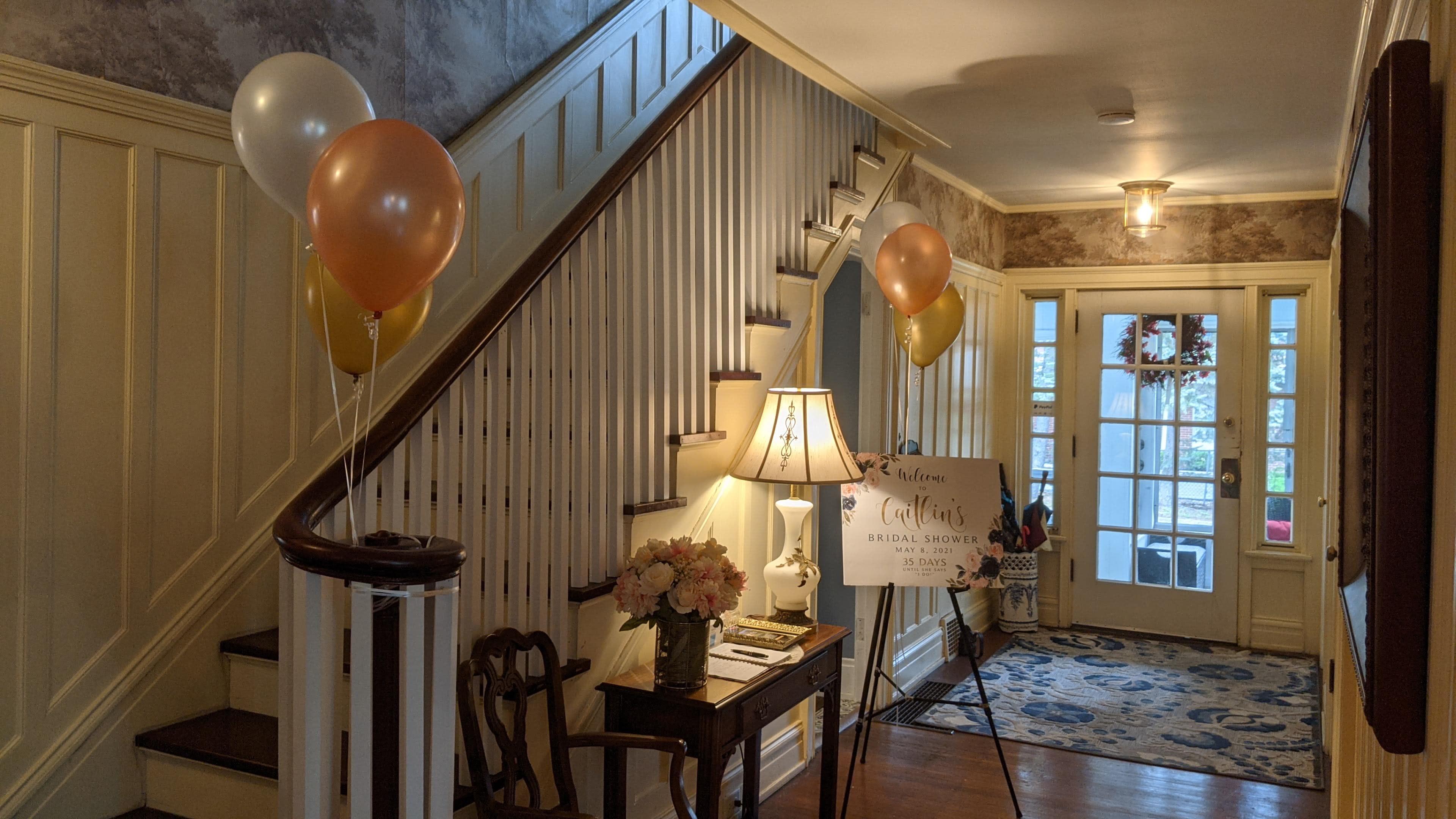 A foyer with a staircase, a table displaying flowers, a sign for a bridal shower, and balloons, leading to a door with natural light.