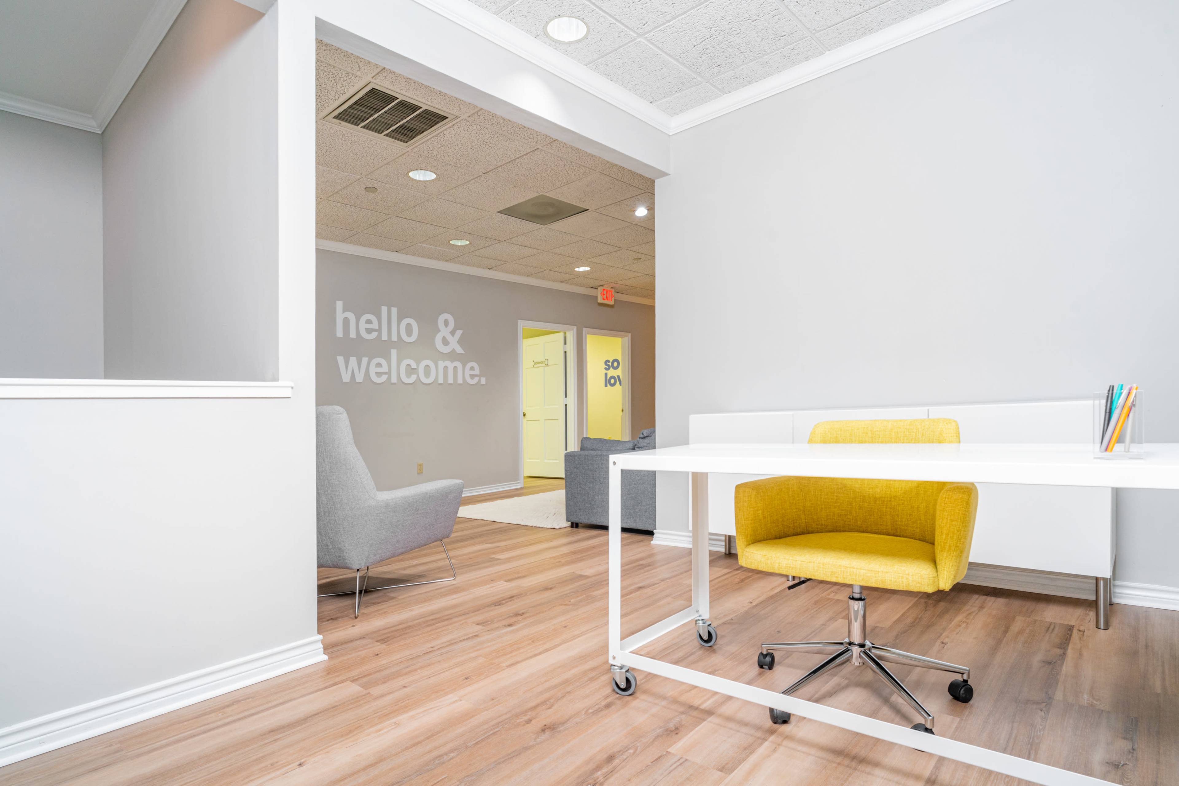 A modern office reception area with a white desk and a yellow chair, leading into a hallway with light-colored walls and welcoming signage.