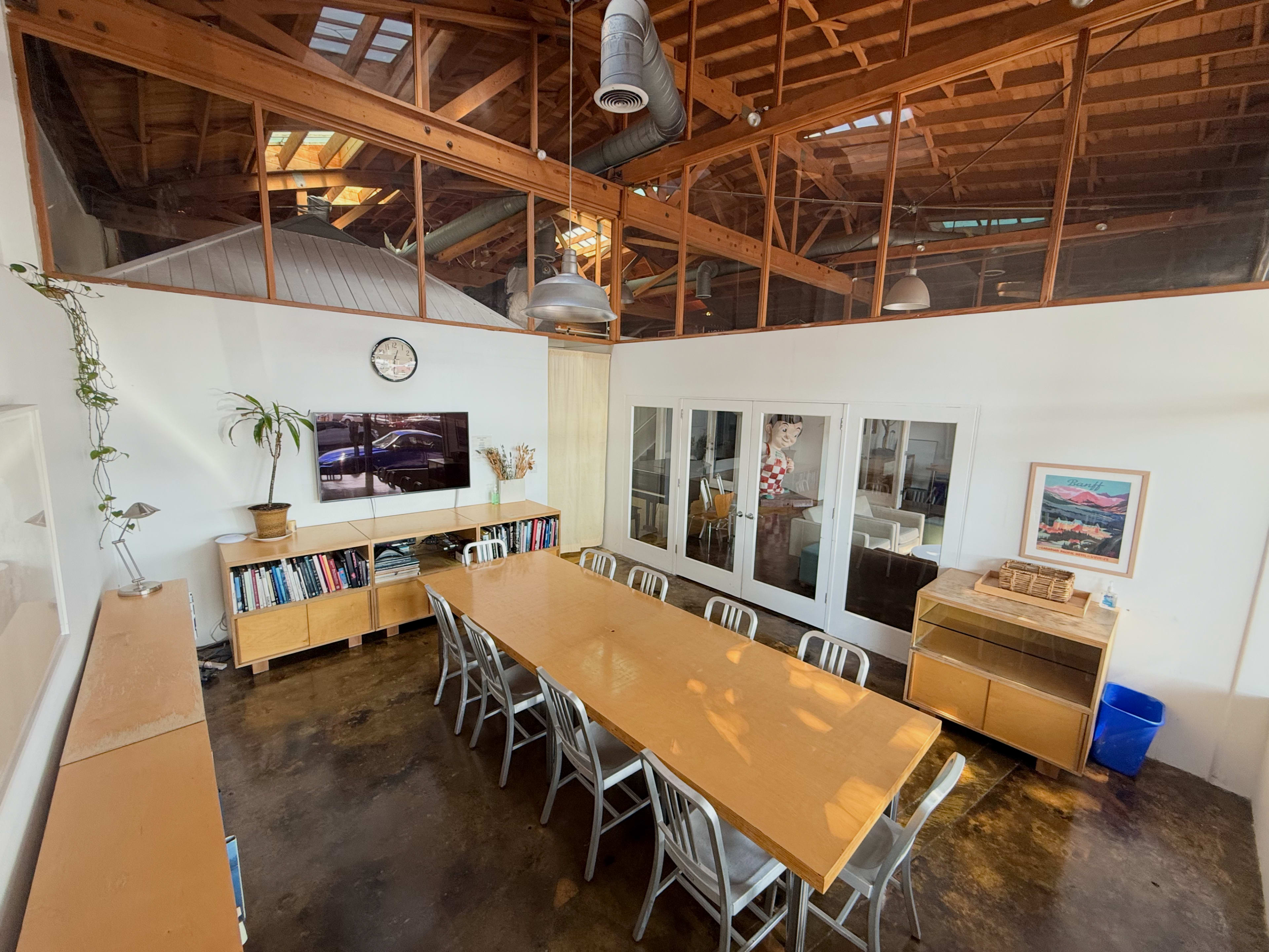 The image shows a spacious meeting room with a long wooden table, metal chairs, large windows, and a television mounted on the wall.