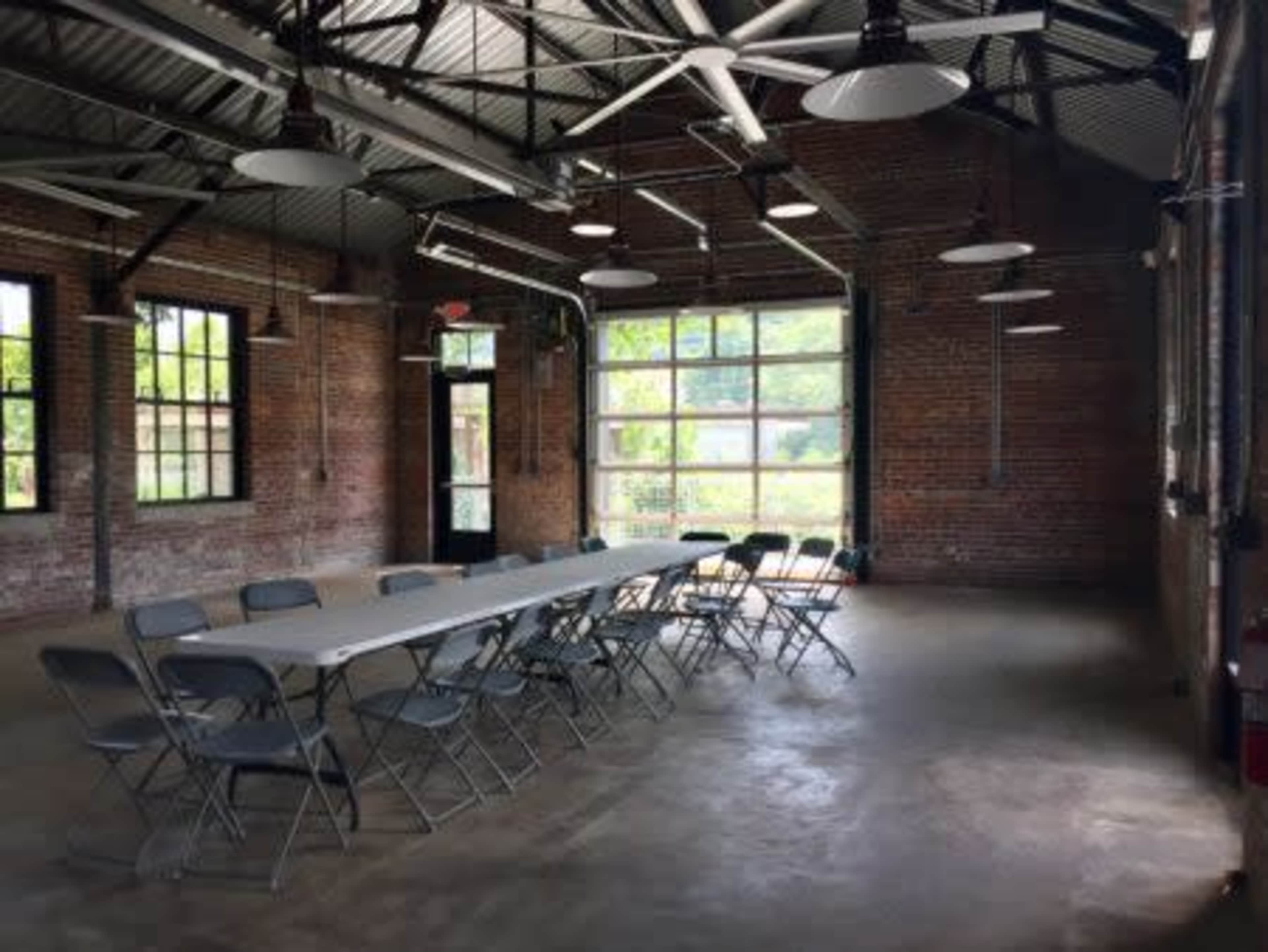 A long table with folding chairs is set up in an industrial-style room featuring exposed brick walls and large windows.