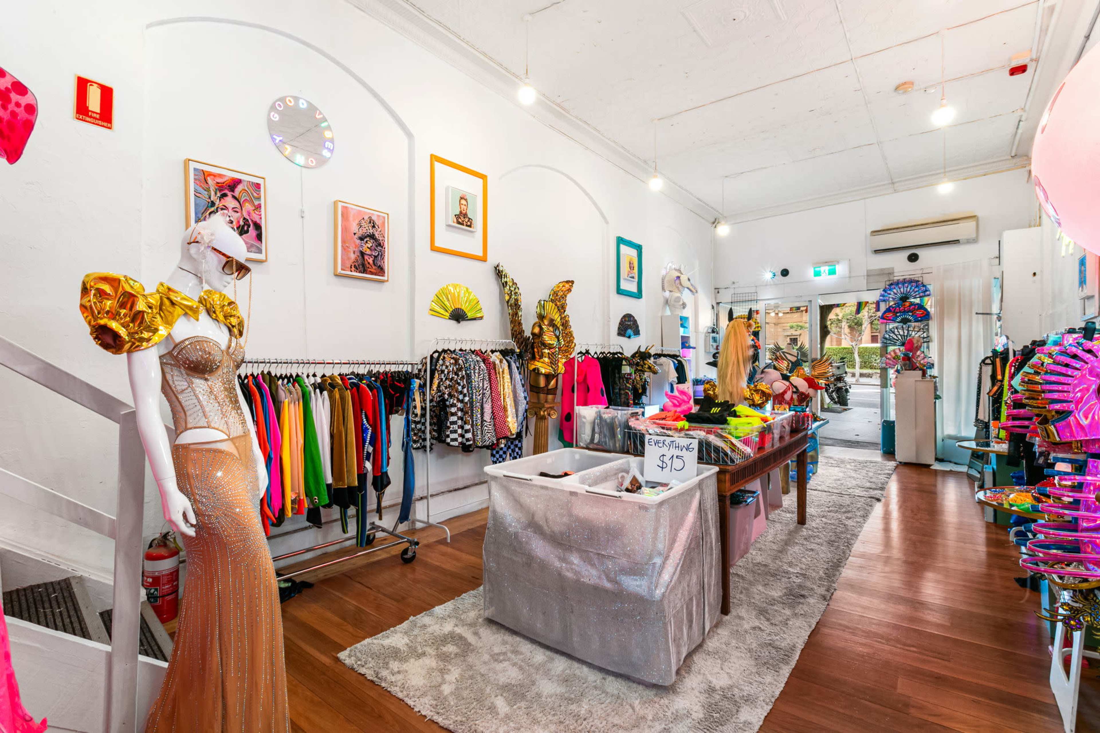 The image shows a vibrant boutique filled with colorful costumes, accessories, and clothing, with mannequins displayed prominently and a central table featuring items for sale.