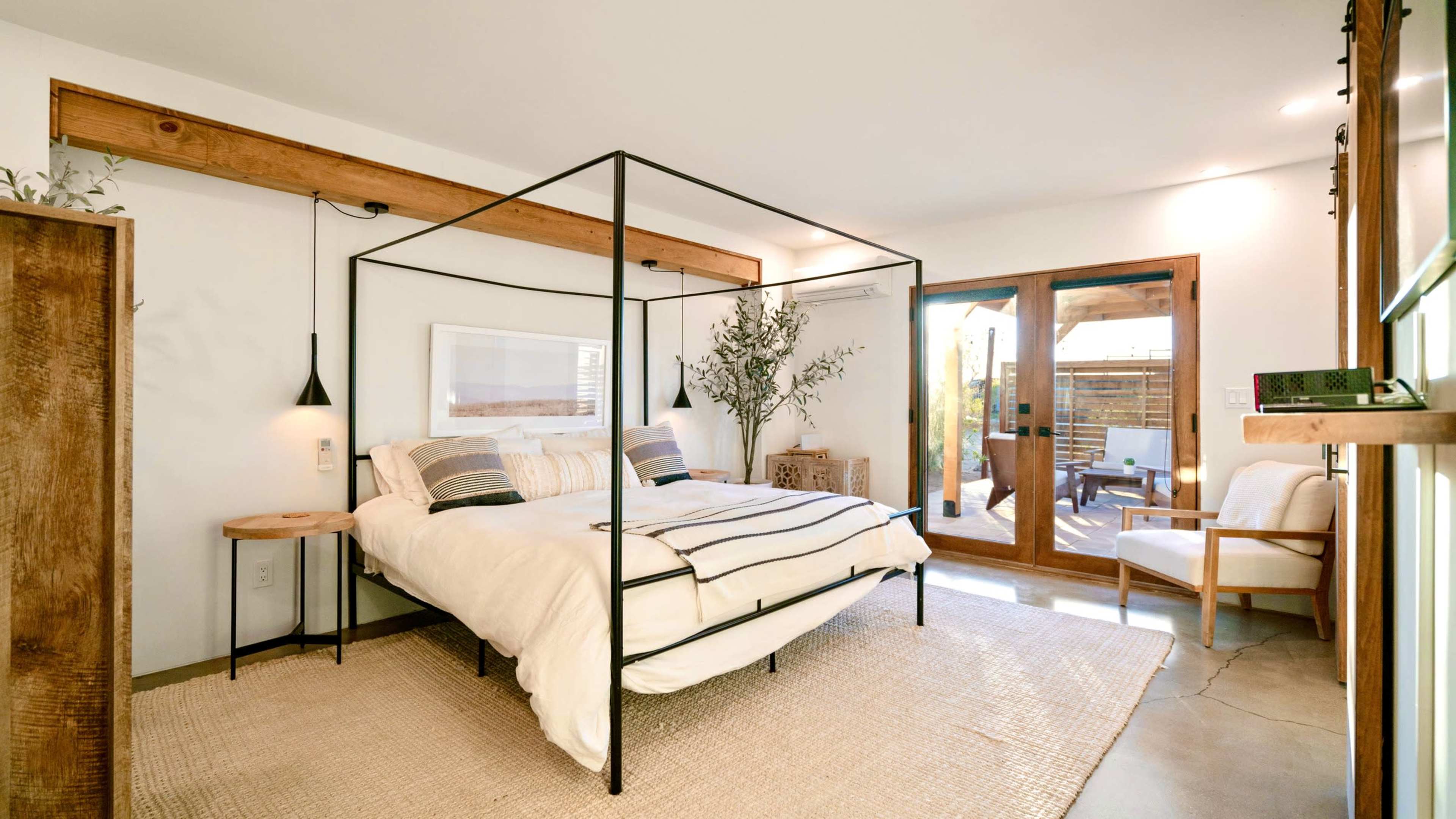 The image shows a modern bedroom featuring a four-poster bed, minimalistic decor, and large glass doors that lead to an outdoor space.