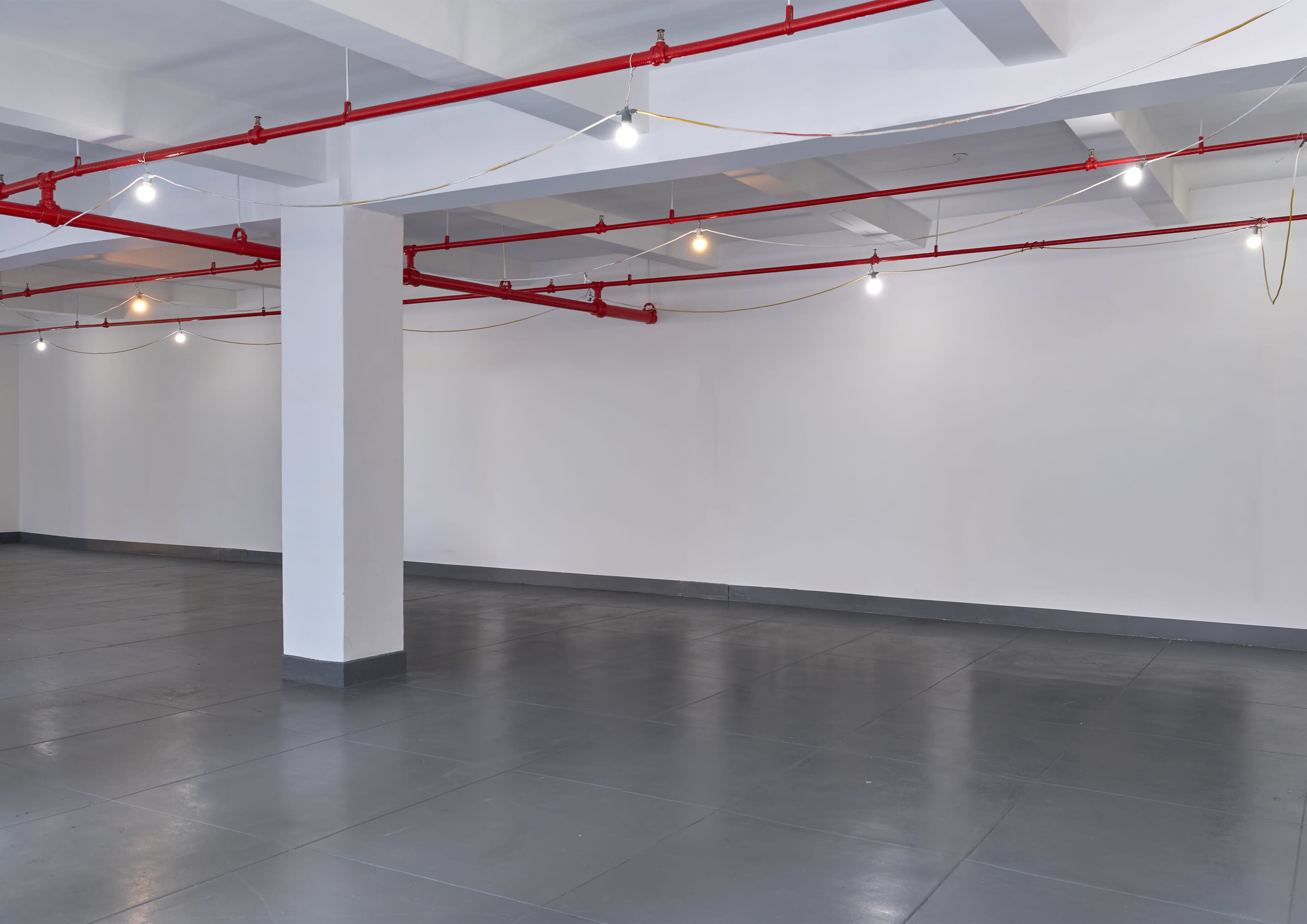 The image shows an empty, minimalist interior space with gray flooring, white walls, and exposed red piping along the ceiling.