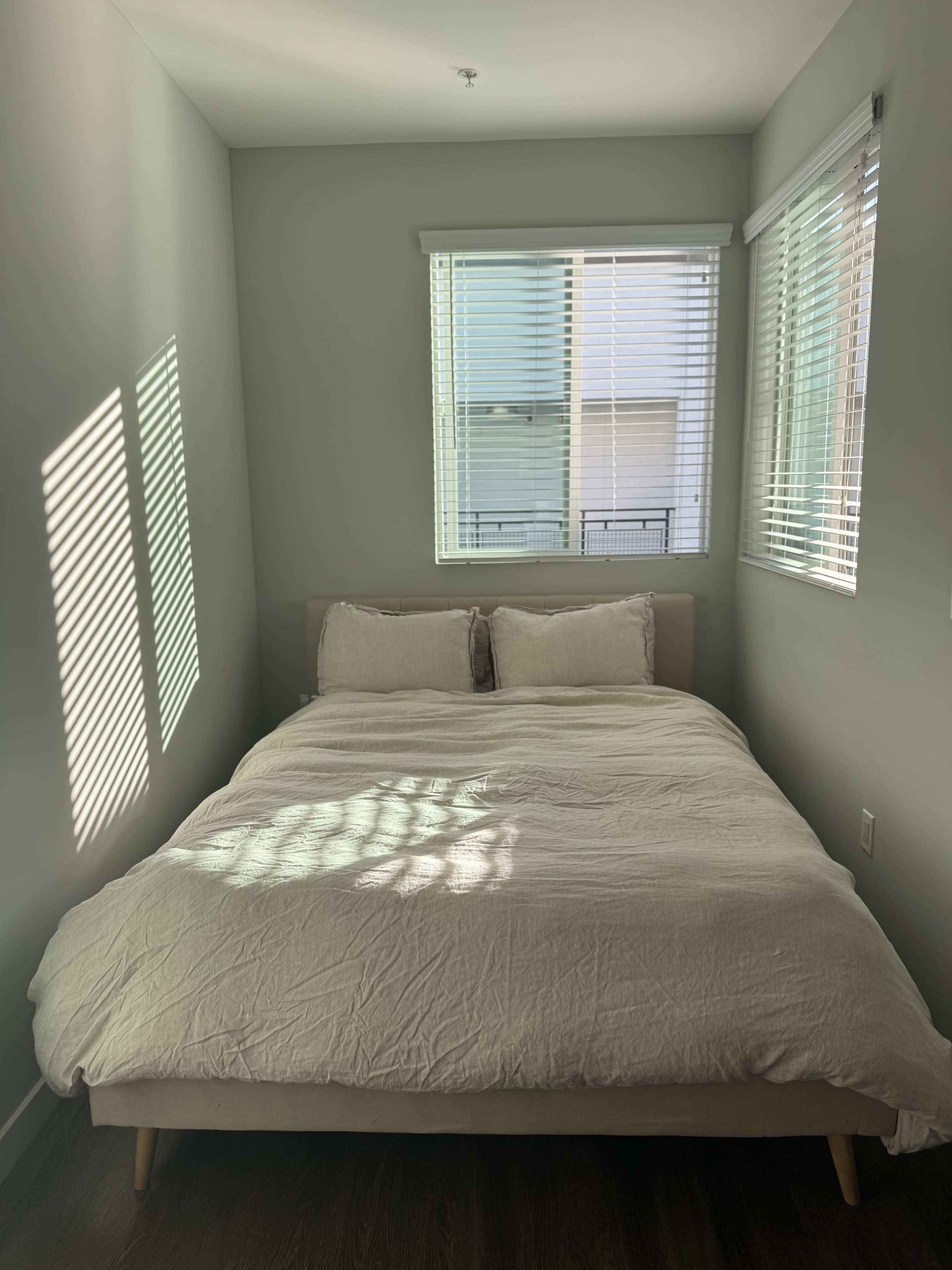 A bed with a light-colored comforter is positioned in a room with two windows, allowing sunlight to cast shadows on the wall.
