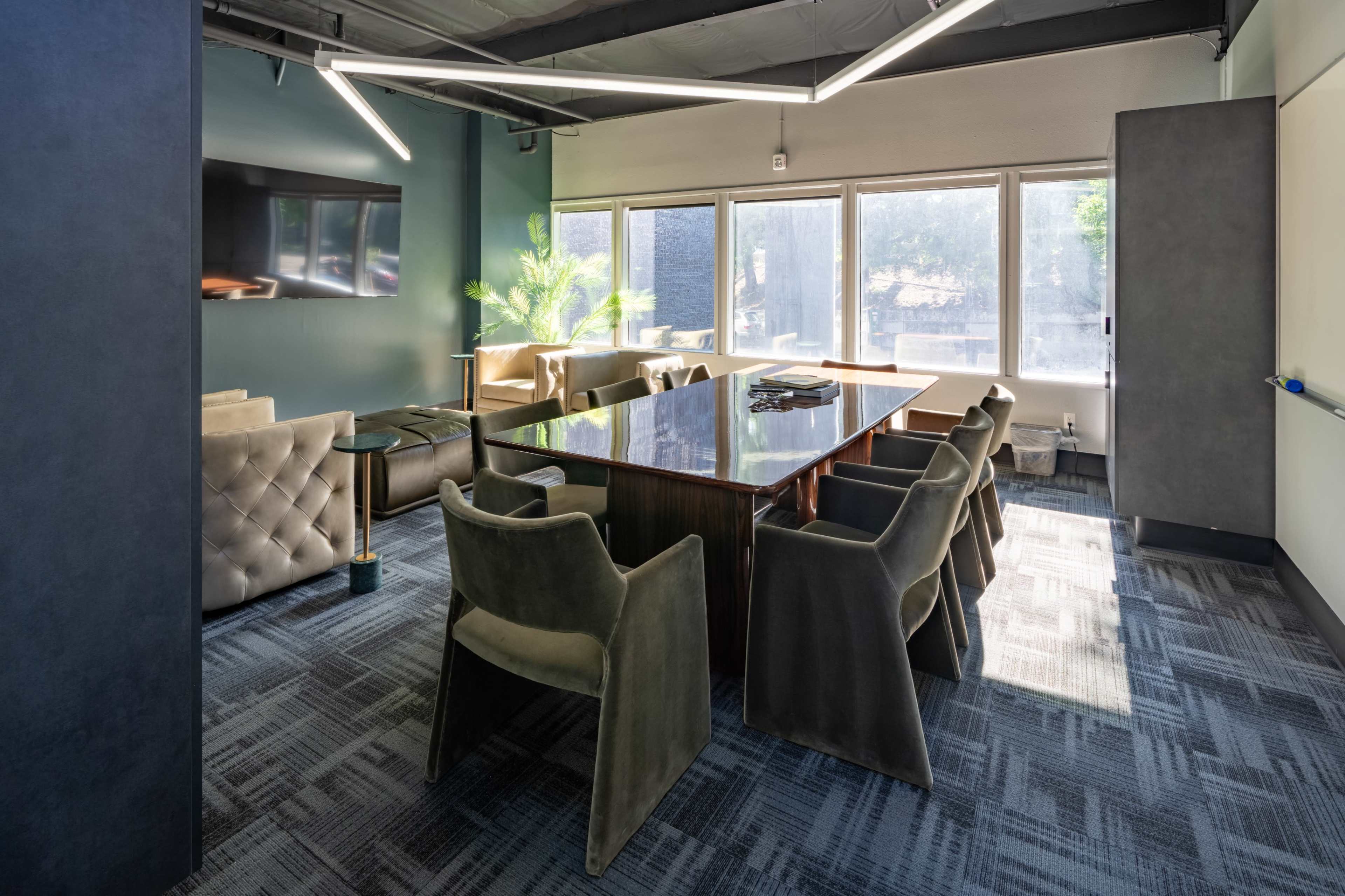 A modern conference room features a large table surrounded by upholstered chairs and large windows providing natural light.