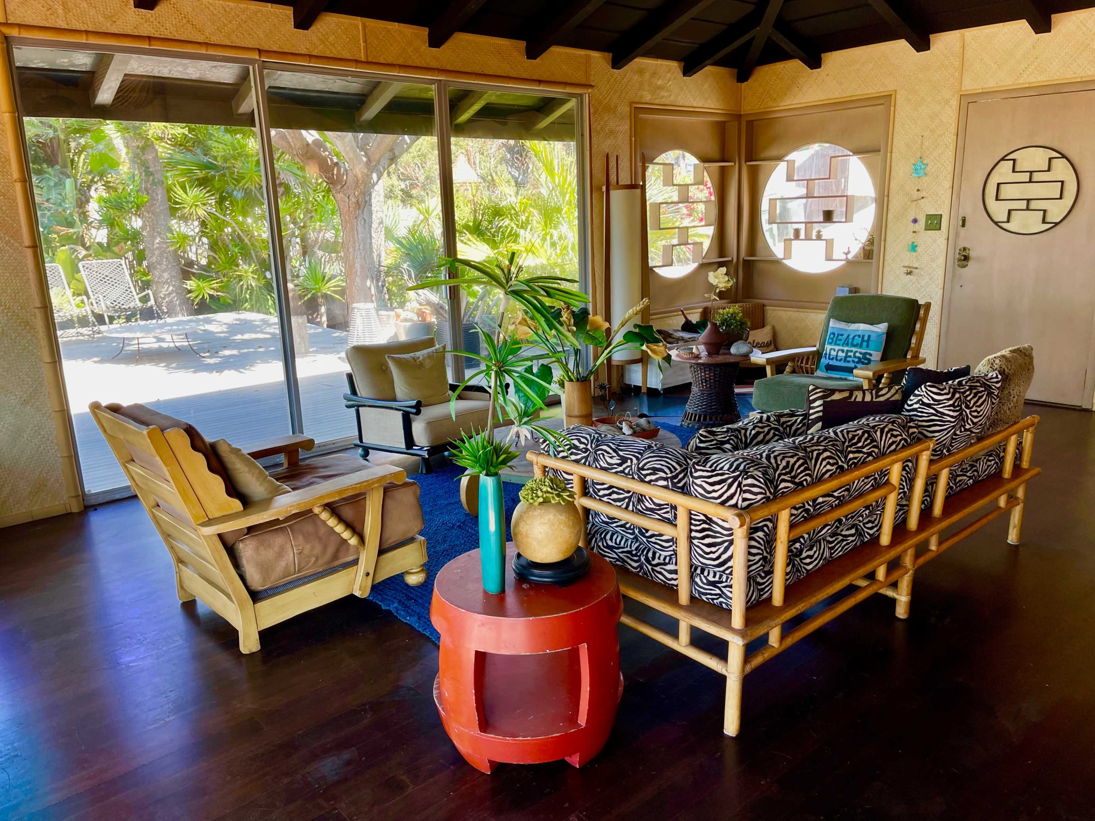 A spacious living room with bamboo furniture, a large window overlooking a garden, and a rug on a wooden floor.