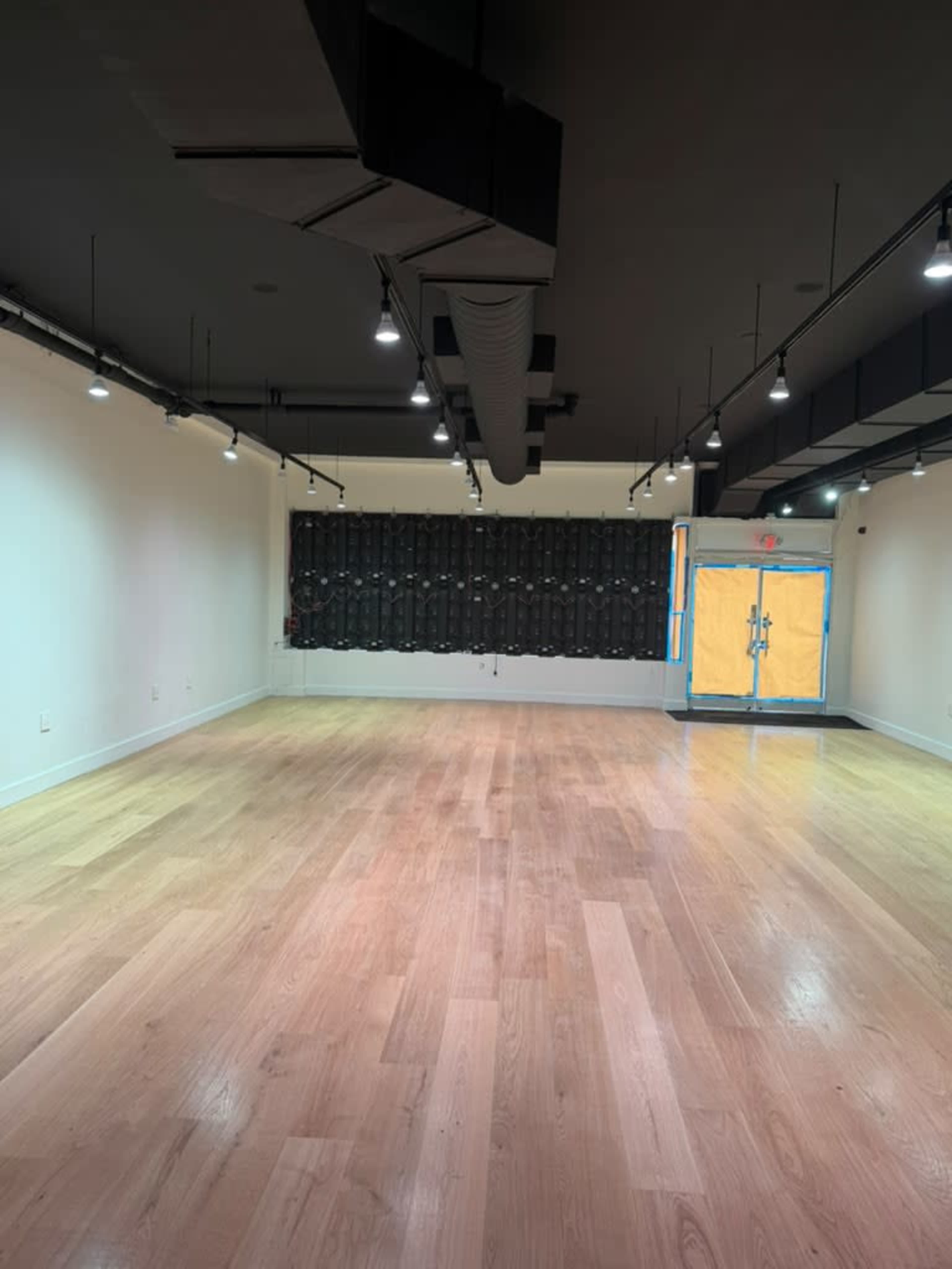 The image shows an empty room with wooden flooring, bare white walls, and a black wall with a door painted in yellow on the right side.