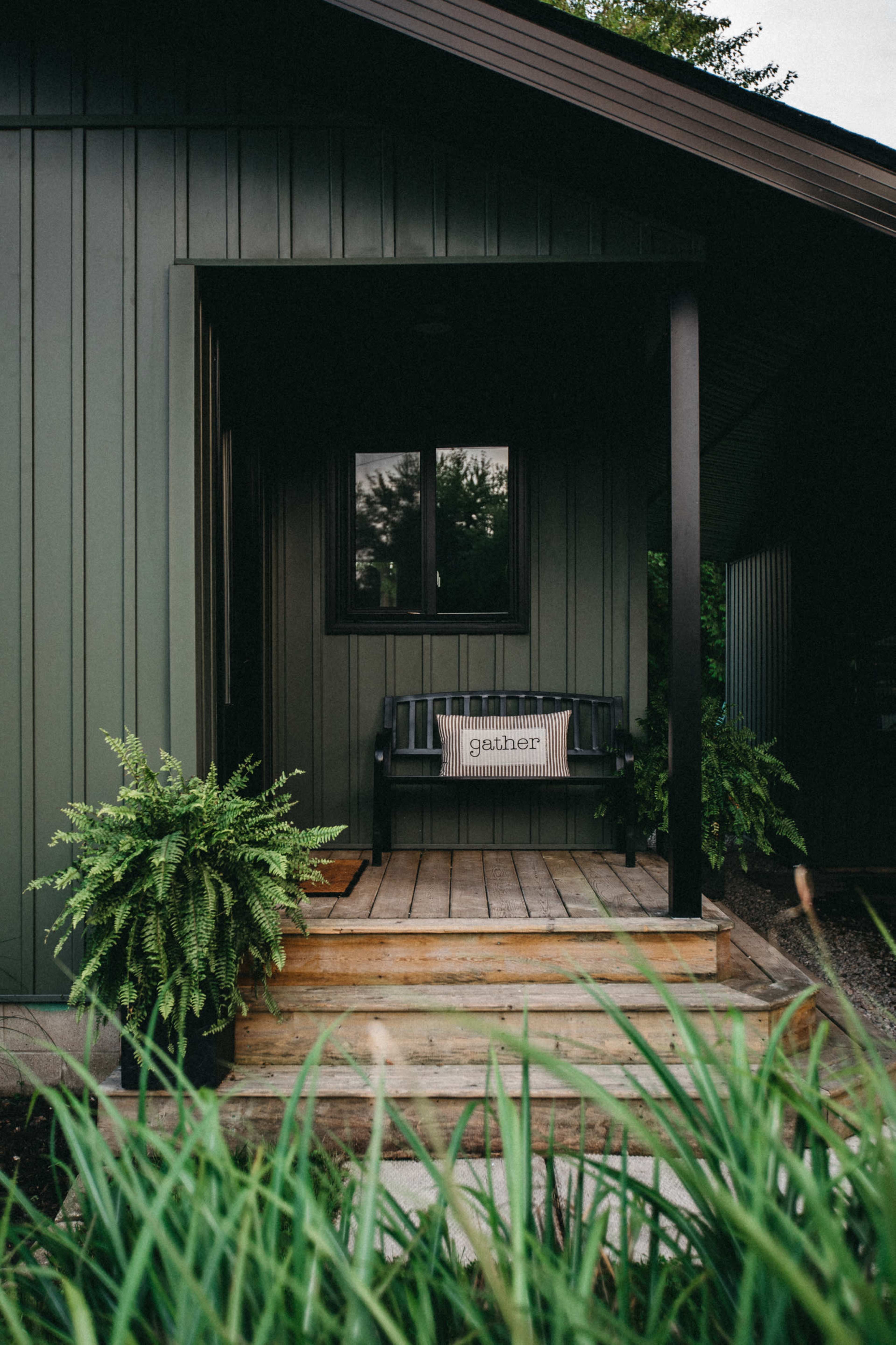 A dark green house features a front porch with a wooden bench and a decorative sign that says "gather."