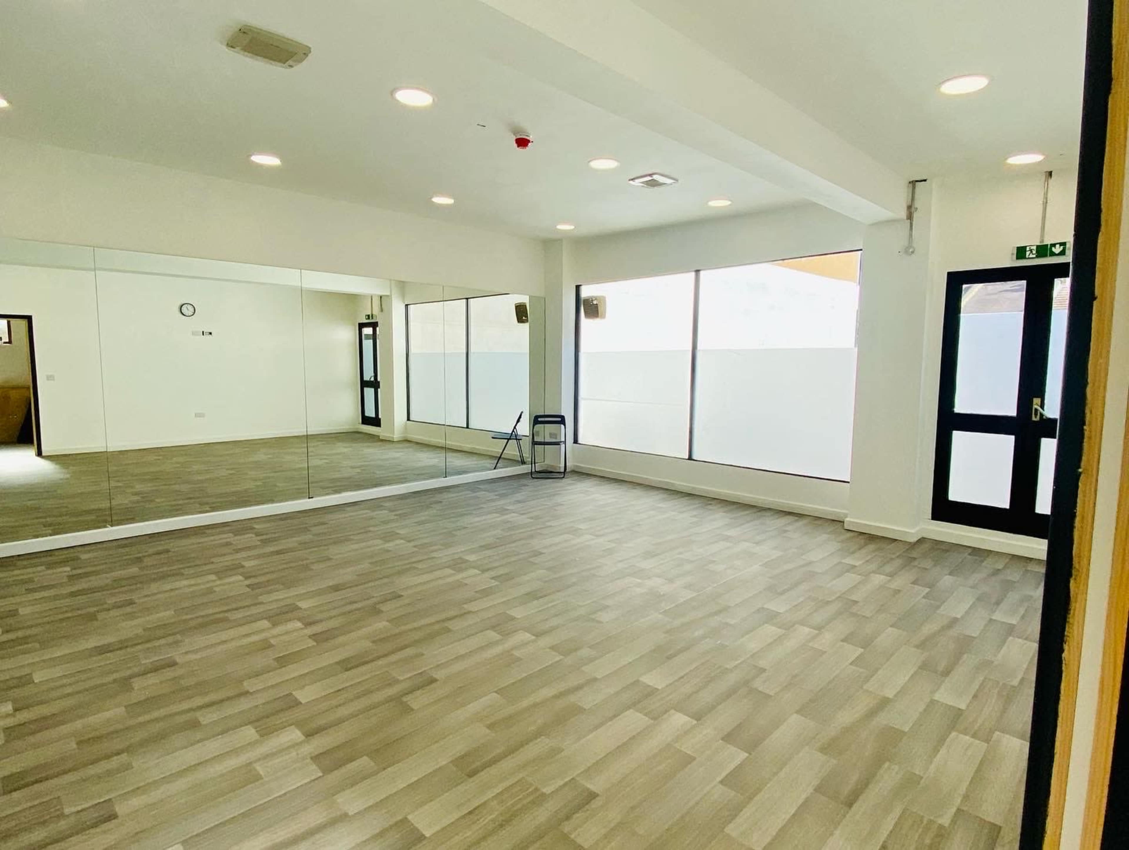 An empty room with wooden flooring, large windows, and a wall of mirrors.