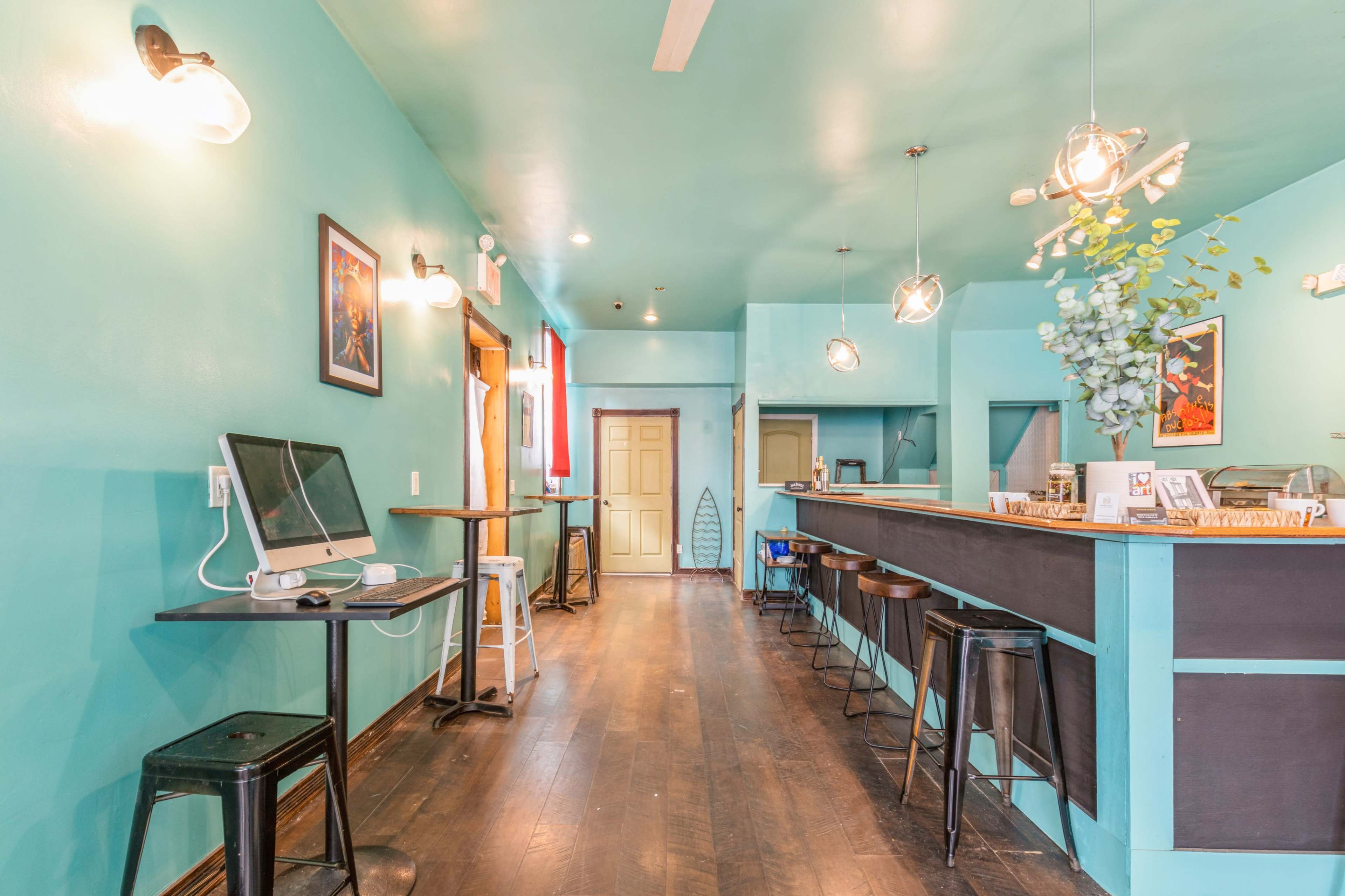 The image shows a brightly colored café interior with teal walls, a bar area with stools, and a computer setup on a small table.