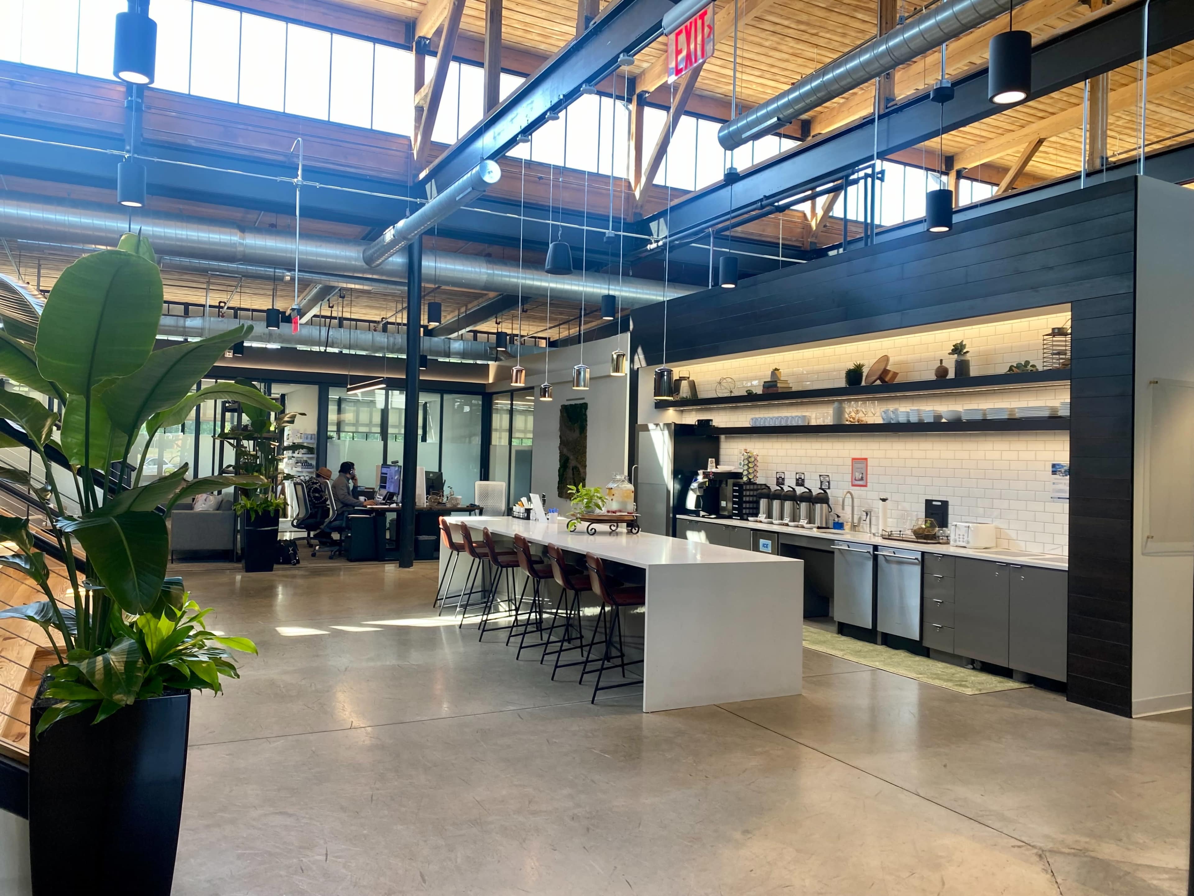 A modern open office space featuring a kitchen area with a countertop, coffee machines, and seating, alongside workstations with people in the background.