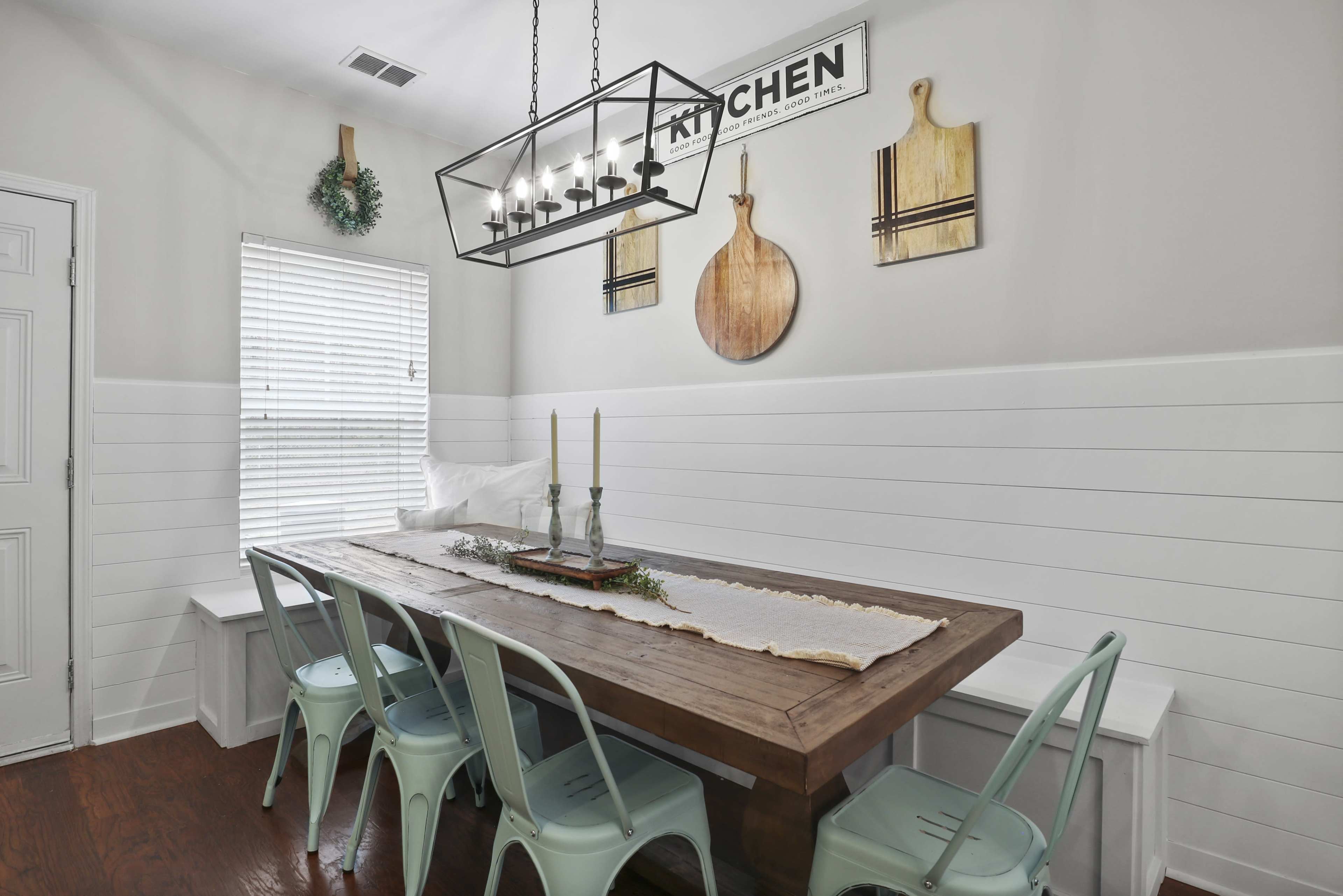 A dining area features a wooden table surrounded by light-colored chairs, with a chandelier above and a window adorned with blinds.