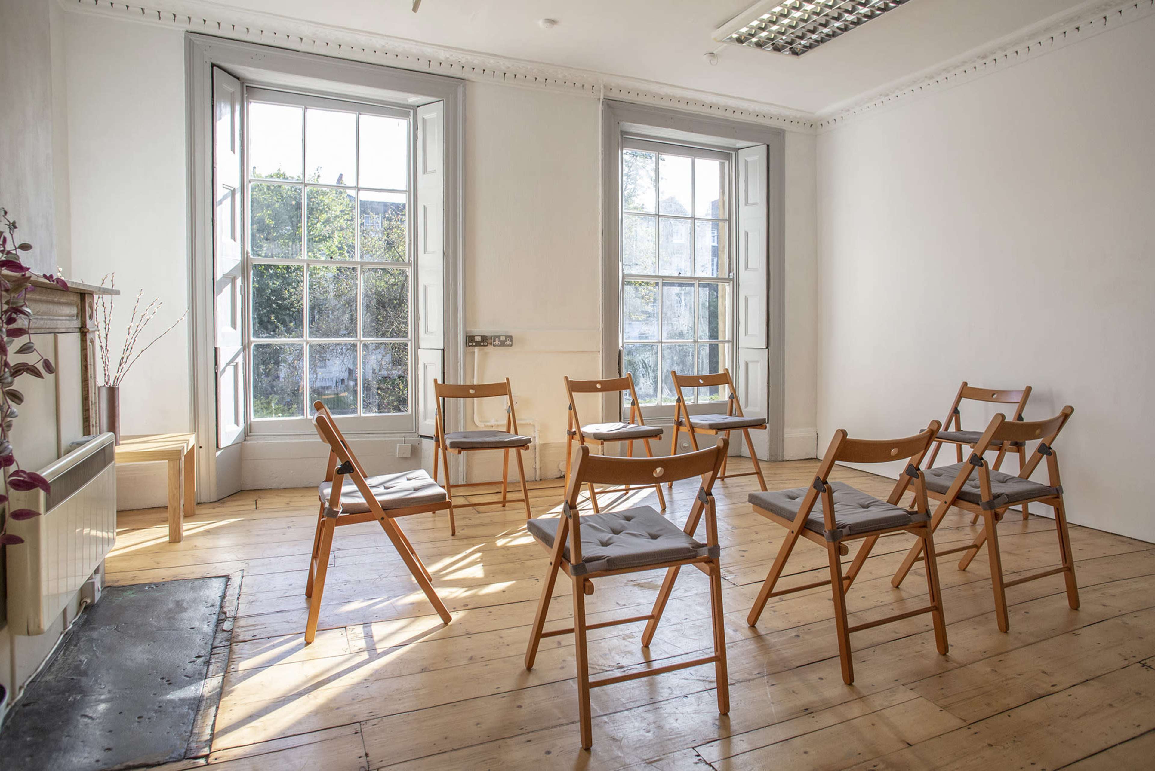 A round arrangement of wooden chairs is set up in a bright, minimally furnished room with large windows.