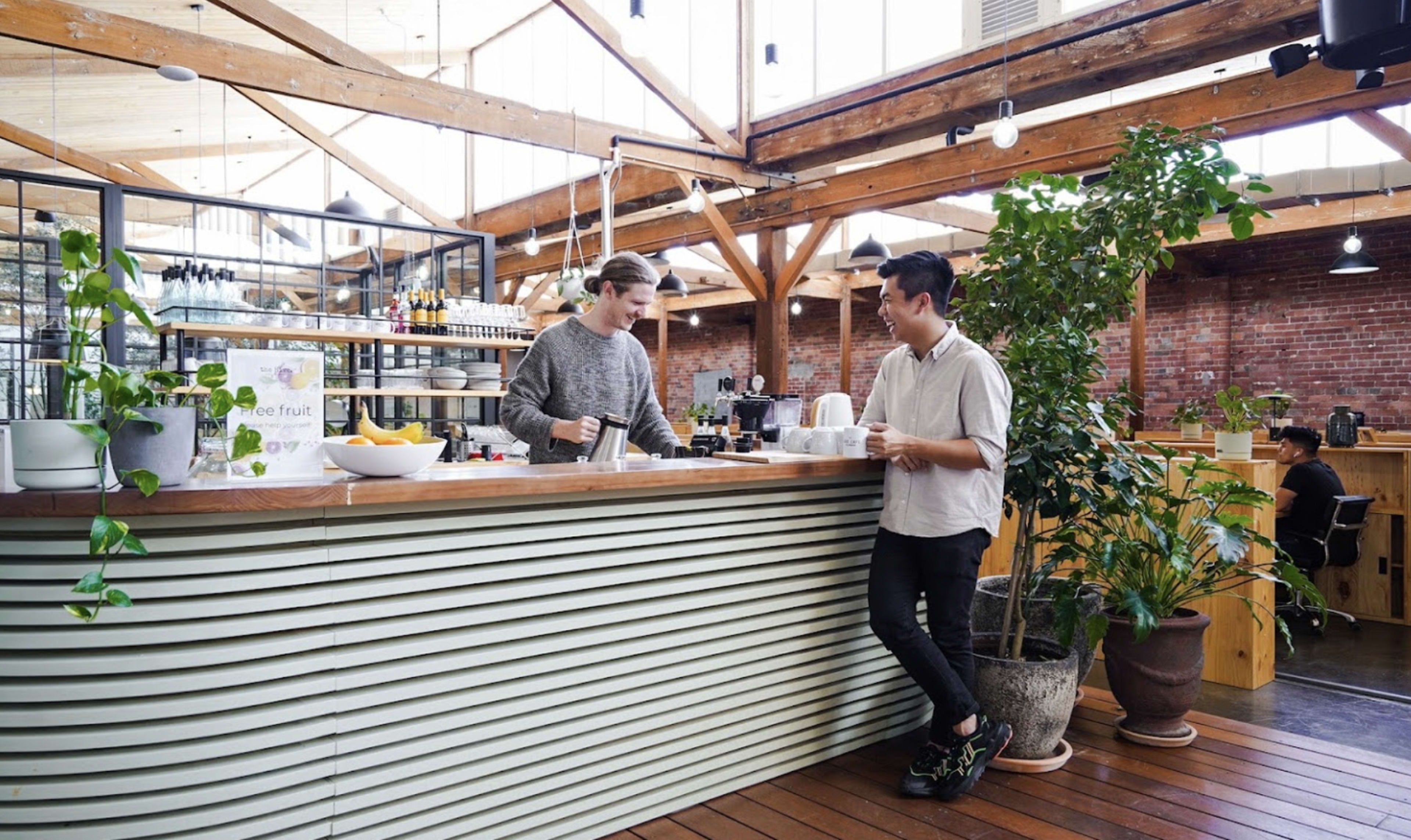 Two individuals are conversing at a wooden counter in a modern cafe with exposed brick walls and indoor plants.