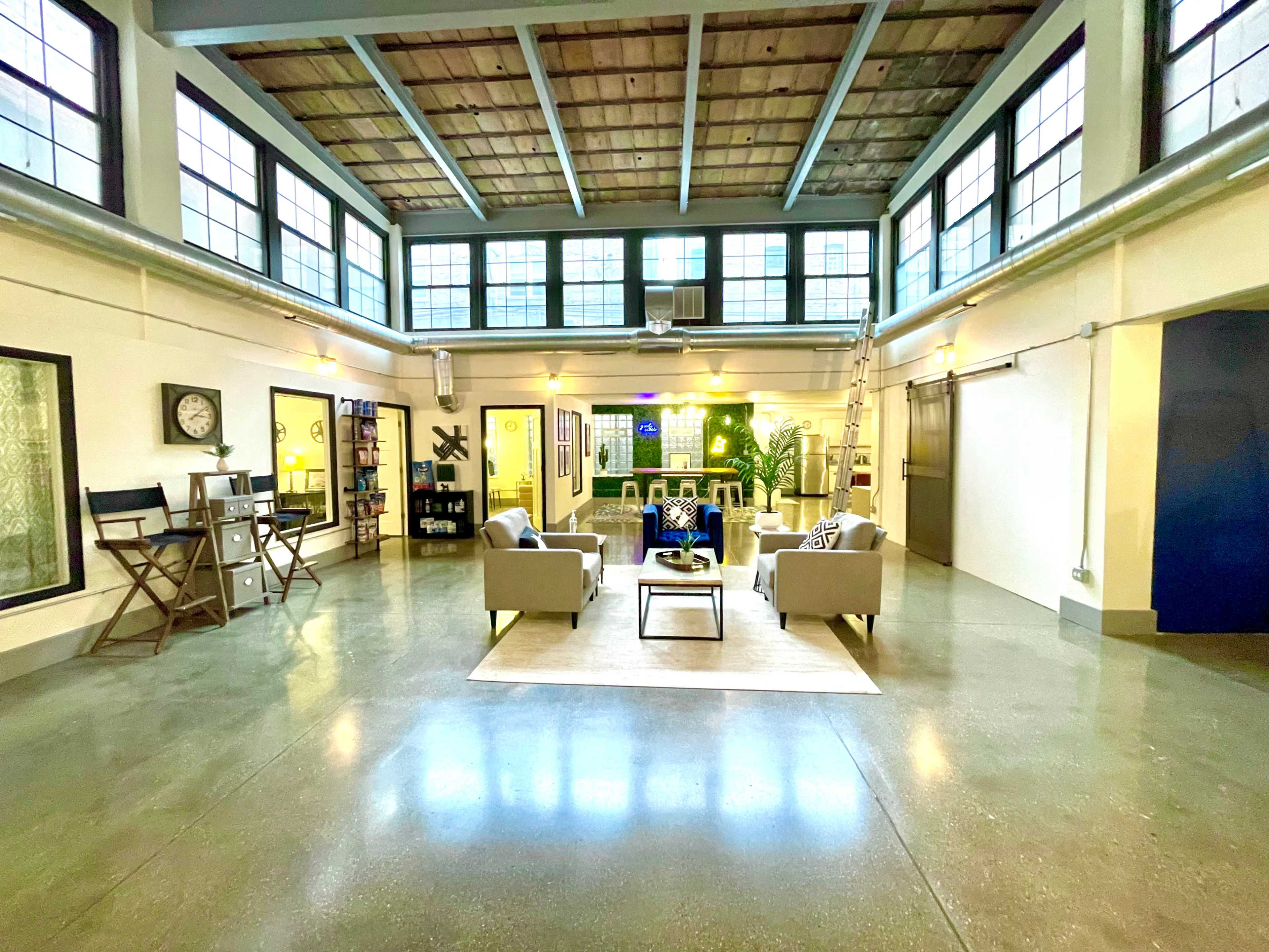 The image shows a spacious, well-lit interior with a seating area featuring two sofas and a coffee table, surrounded by large windows and a polished concrete floor.