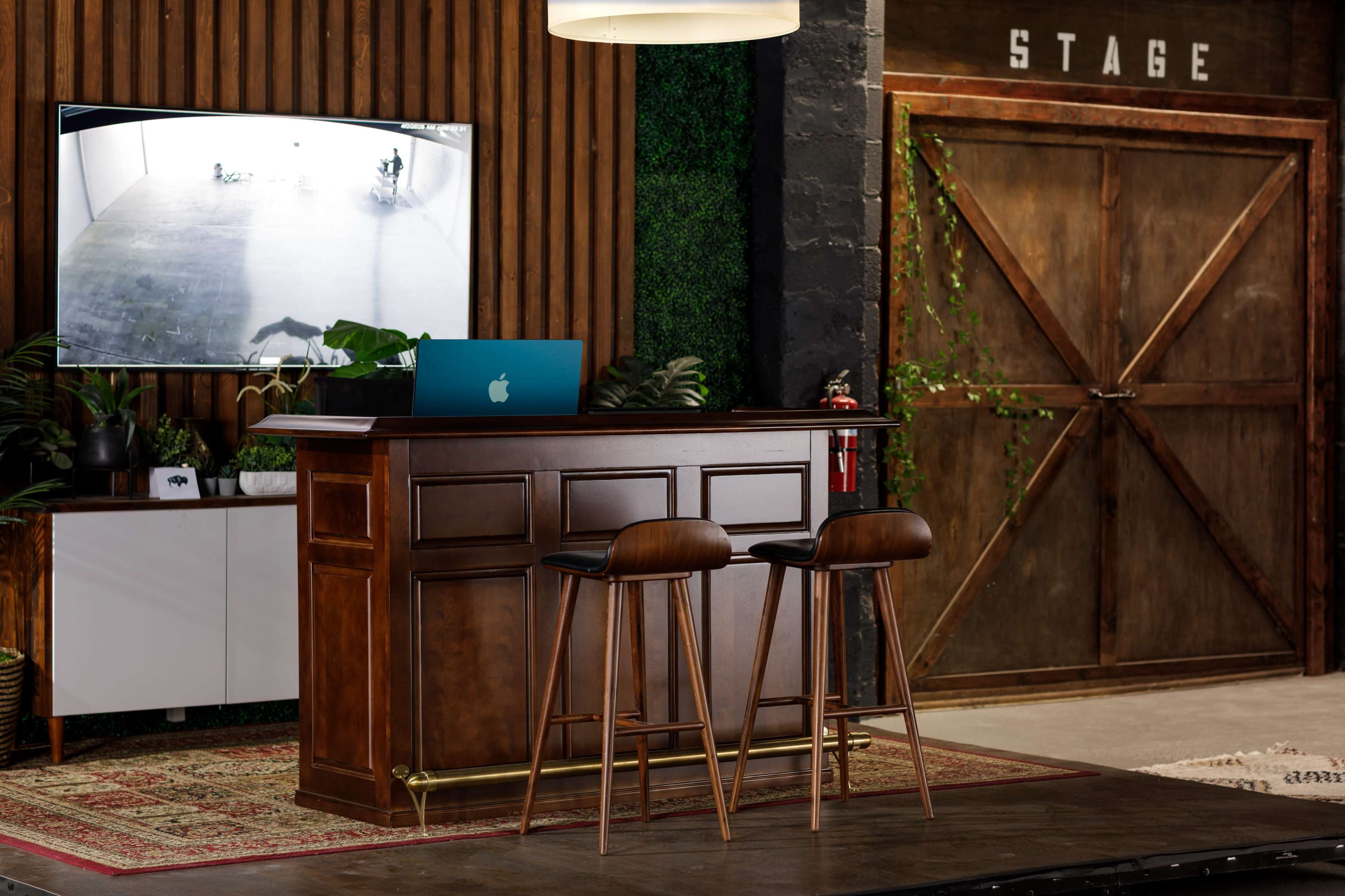 The image depicts a modern bar area with two stools, a wooden counter, a large screen displaying a video feed, and a rustic door labeled "STAGE" in the background.