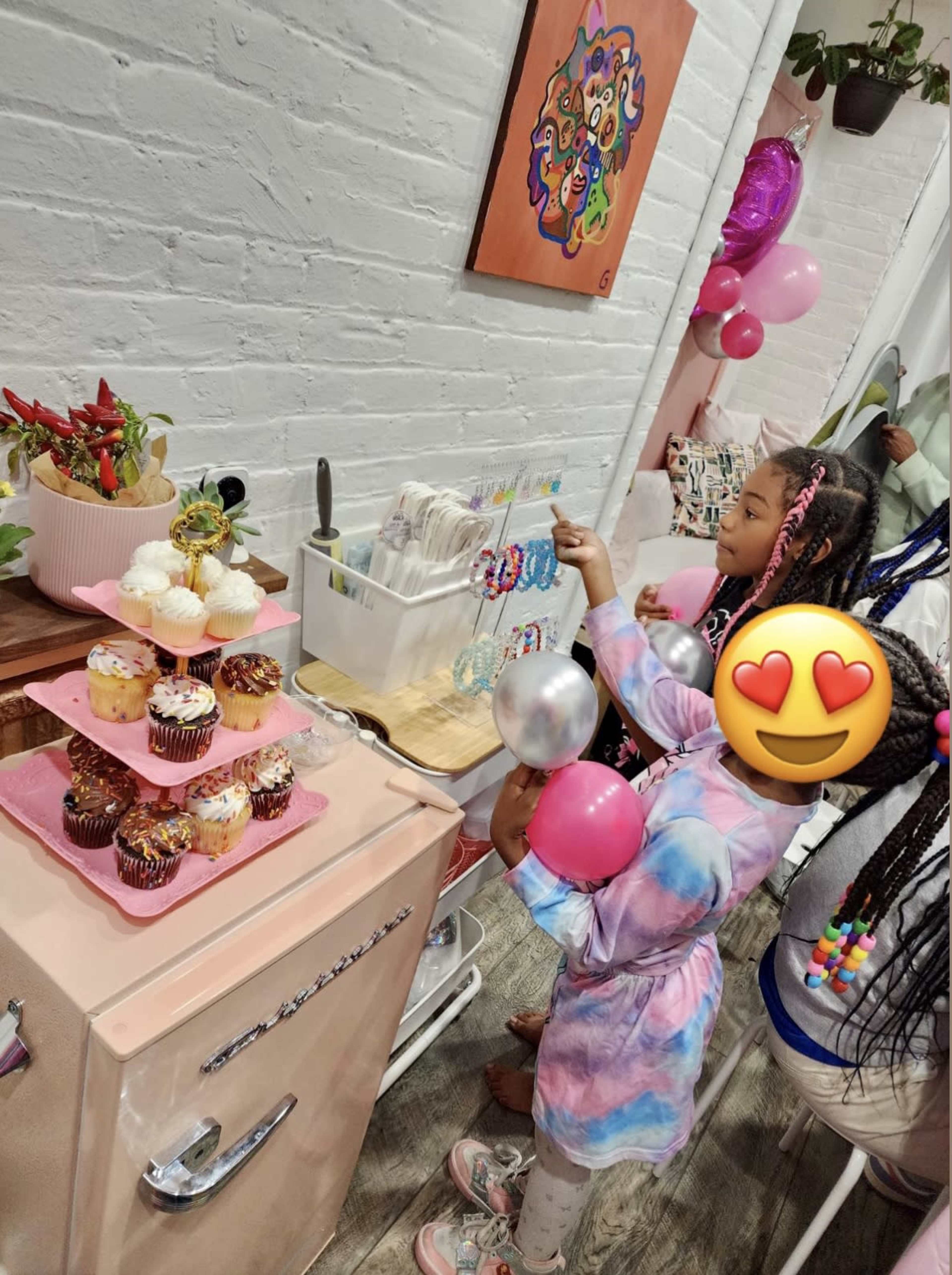 Two children are at a table decorated with cupcakes and balloons, one of them reaching for a treat while the other holds a pink balloon.