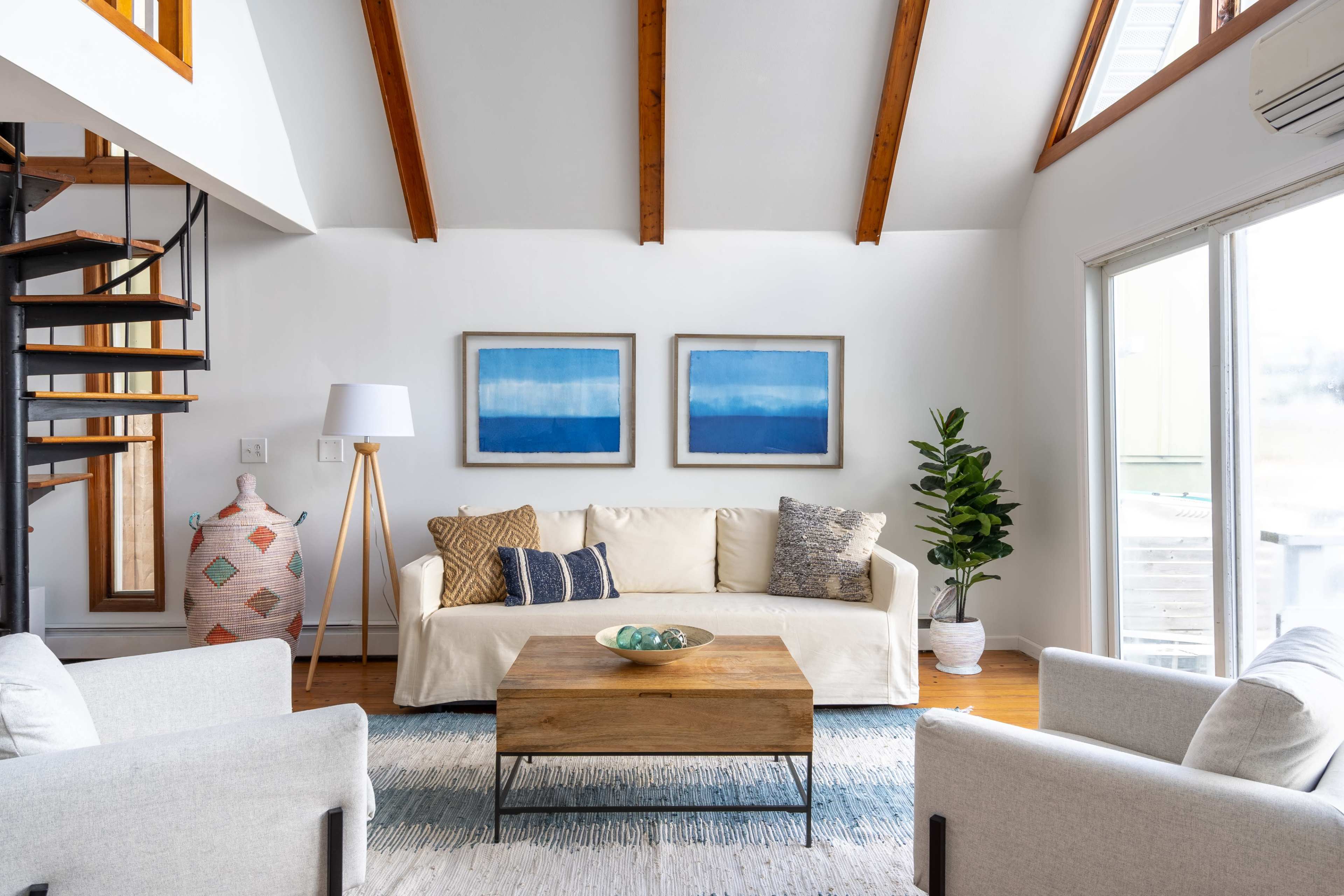 A living room features a light-colored sofa with decorative cushions, a wooden coffee table, two armchairs, and abstract artwork on the walls, alongside a spiral staircase and a large window.