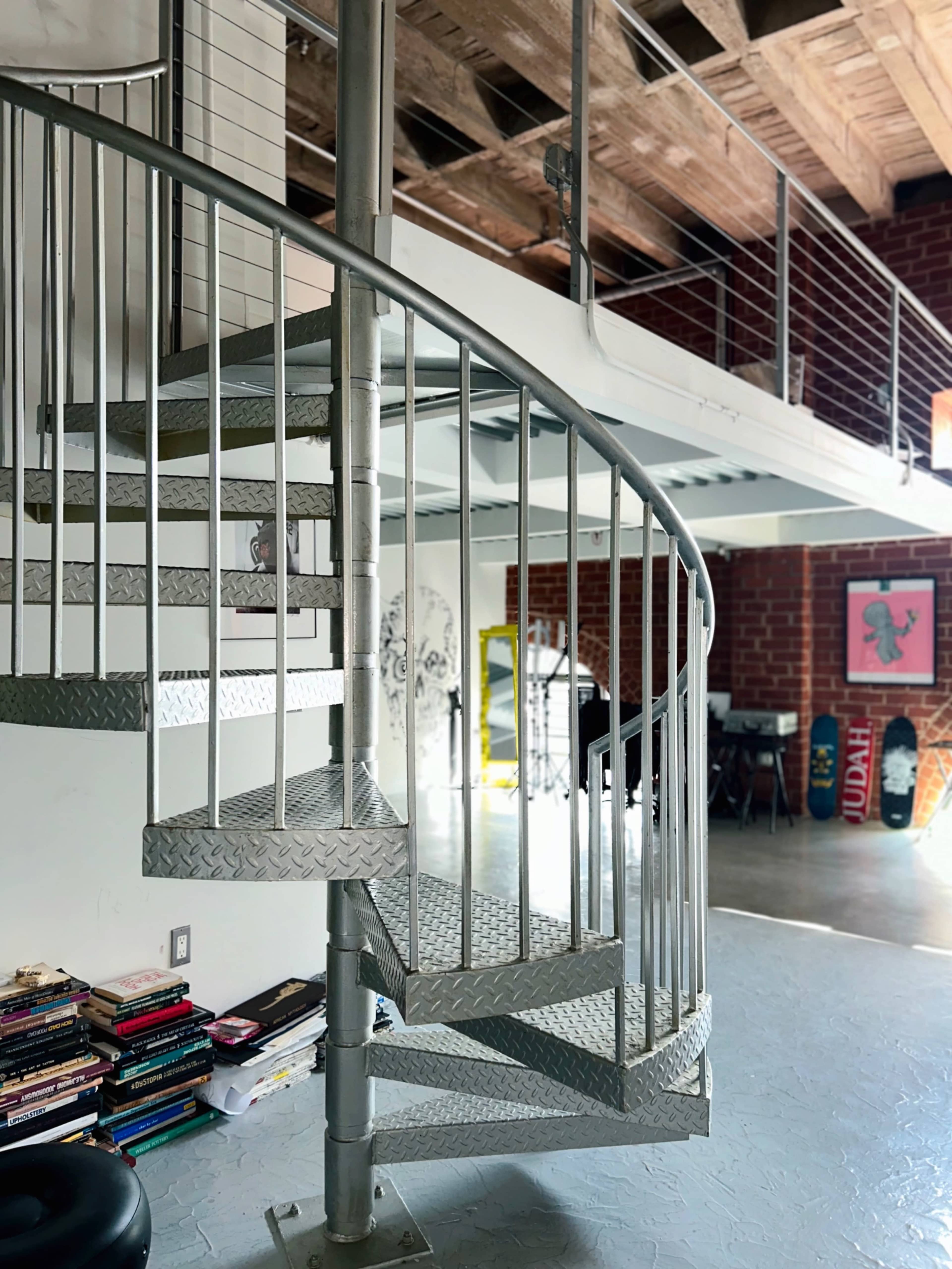 A spiral staircase made of metal leads from a lower level to an upper loft area in a modern open space, with books stacked nearby and artwork hanging on the walls.