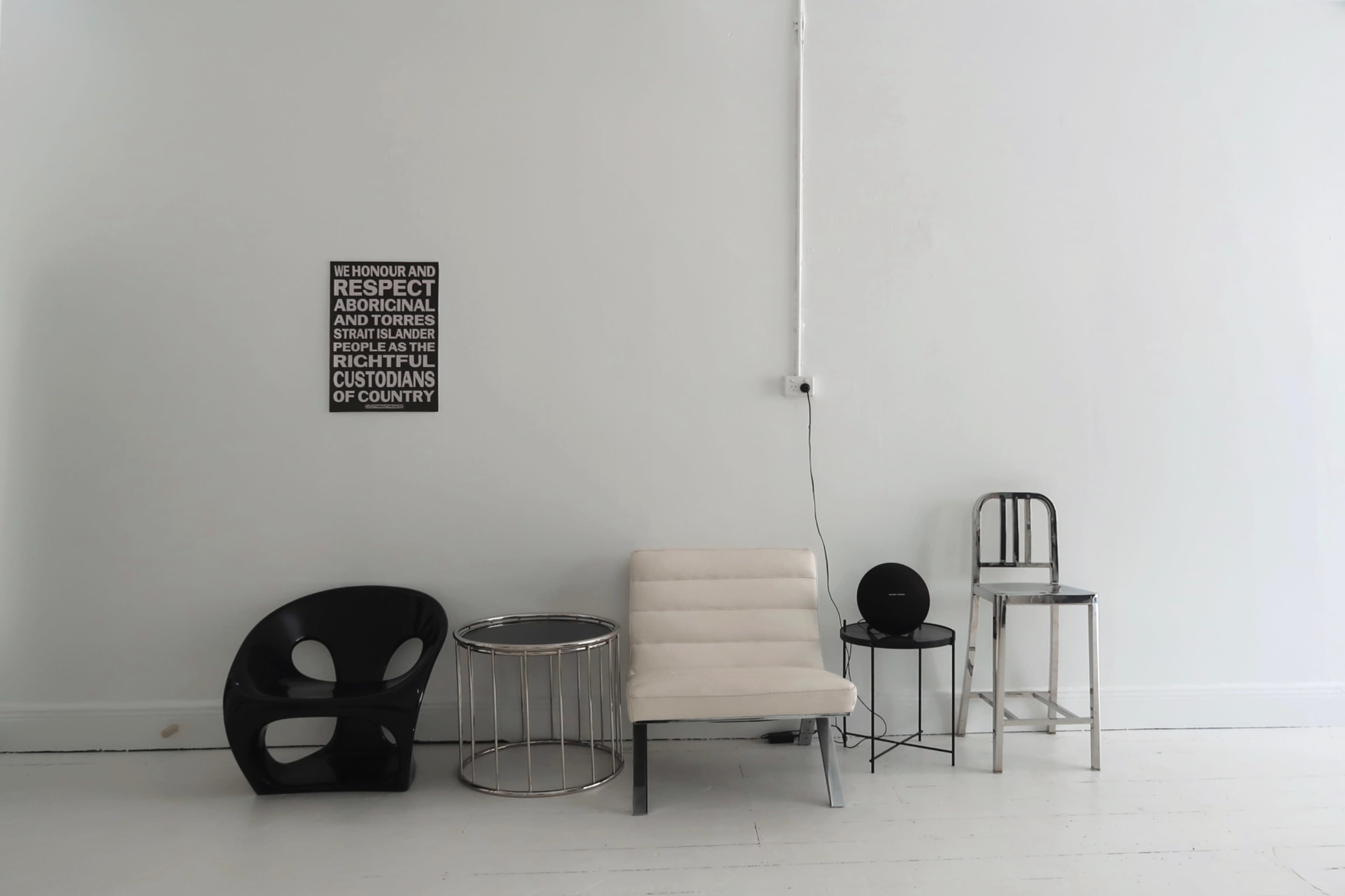 The image shows a minimalist interior featuring a light-colored wall with a sign, a white sofa, a black chair, a metal side table, a round black object, and a wooden stool.