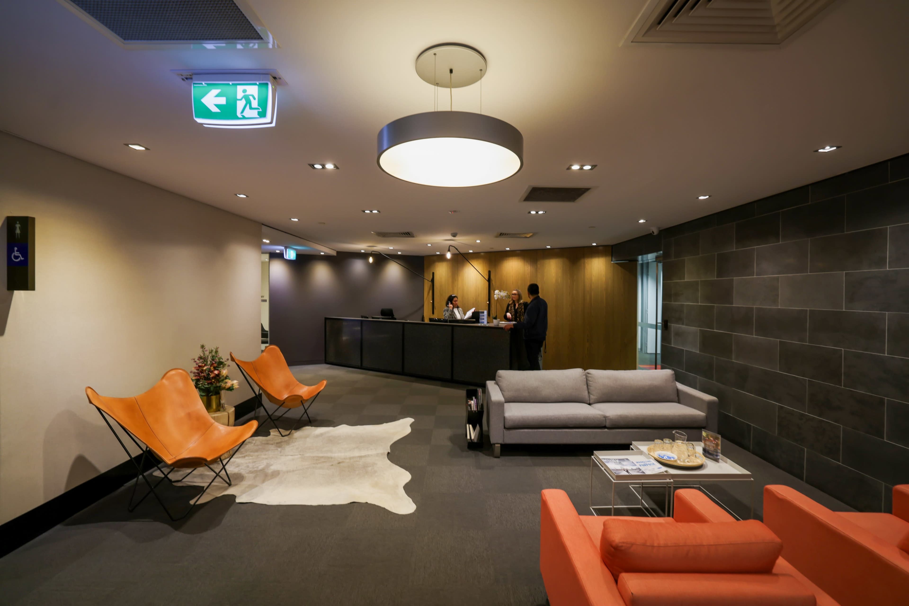 The image shows a modern reception area with a front desk, two orange chairs, a gray sofa, and decorative elements including a cowhide rug.