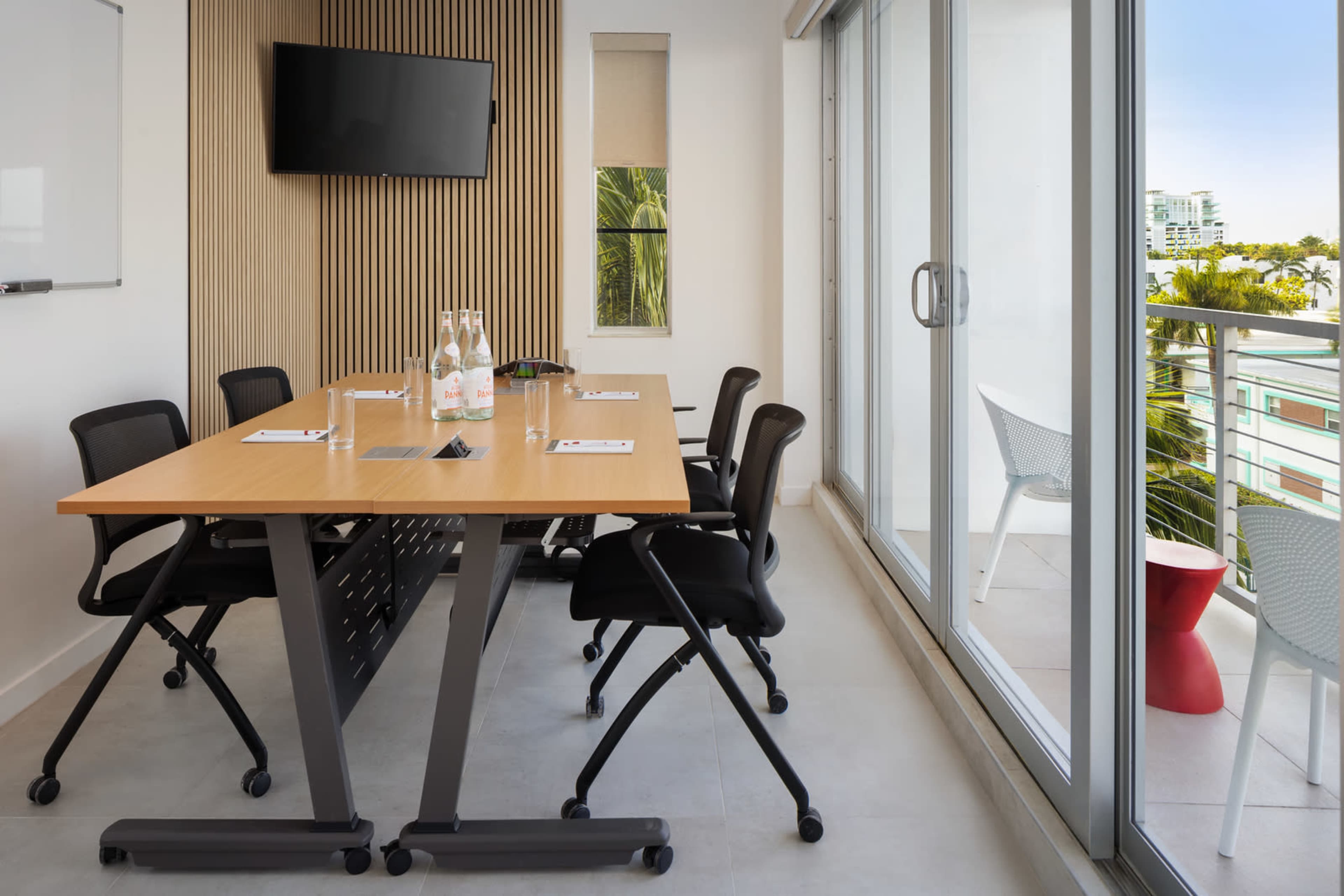 A modern conference room features a wooden table with rolling chairs, a wall-mounted TV, and large windows overlooking greenery.