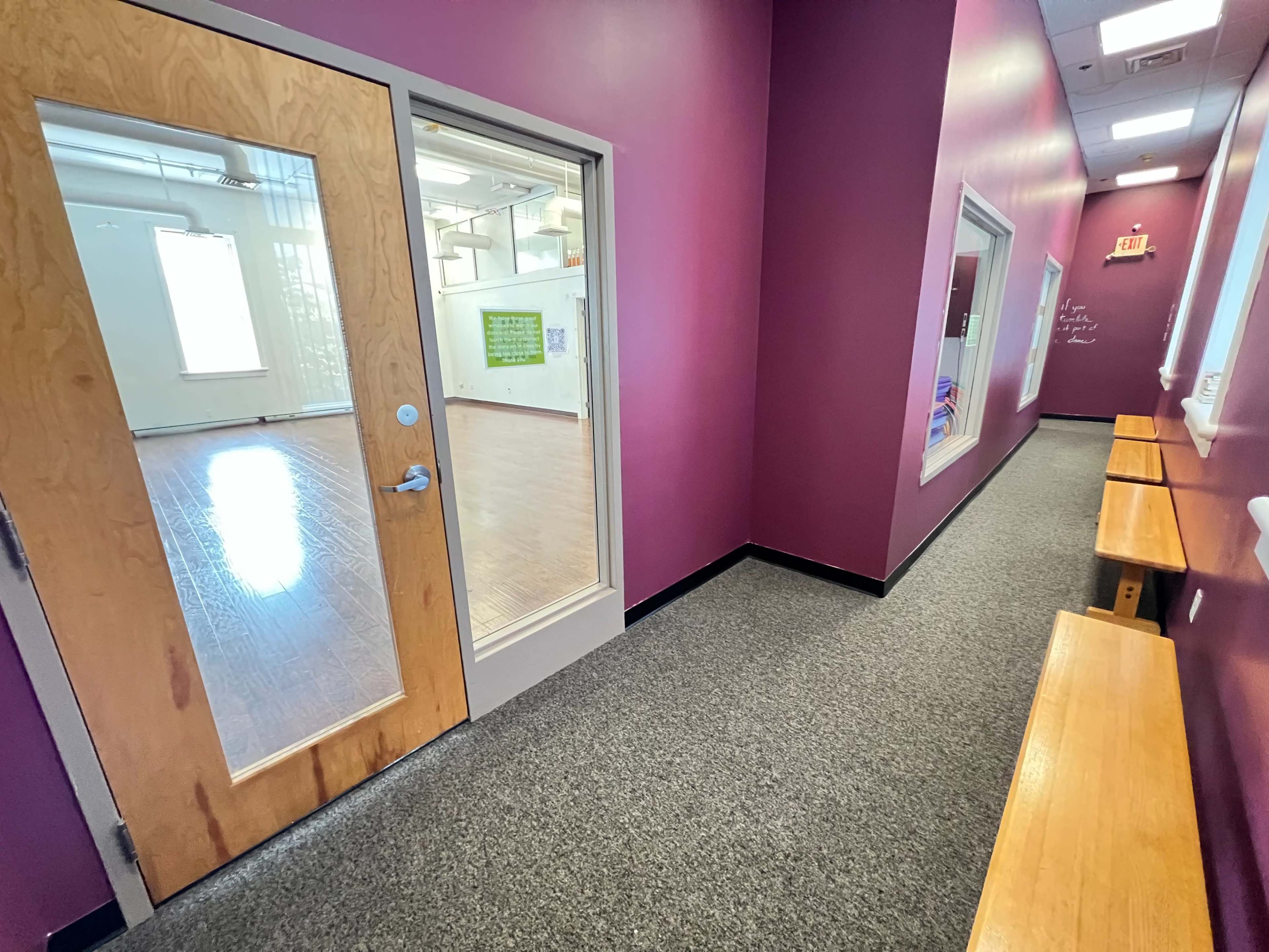 A narrow hallway with a purple wall, a glass door leading to a room with wooden flooring, and two benches along one side.