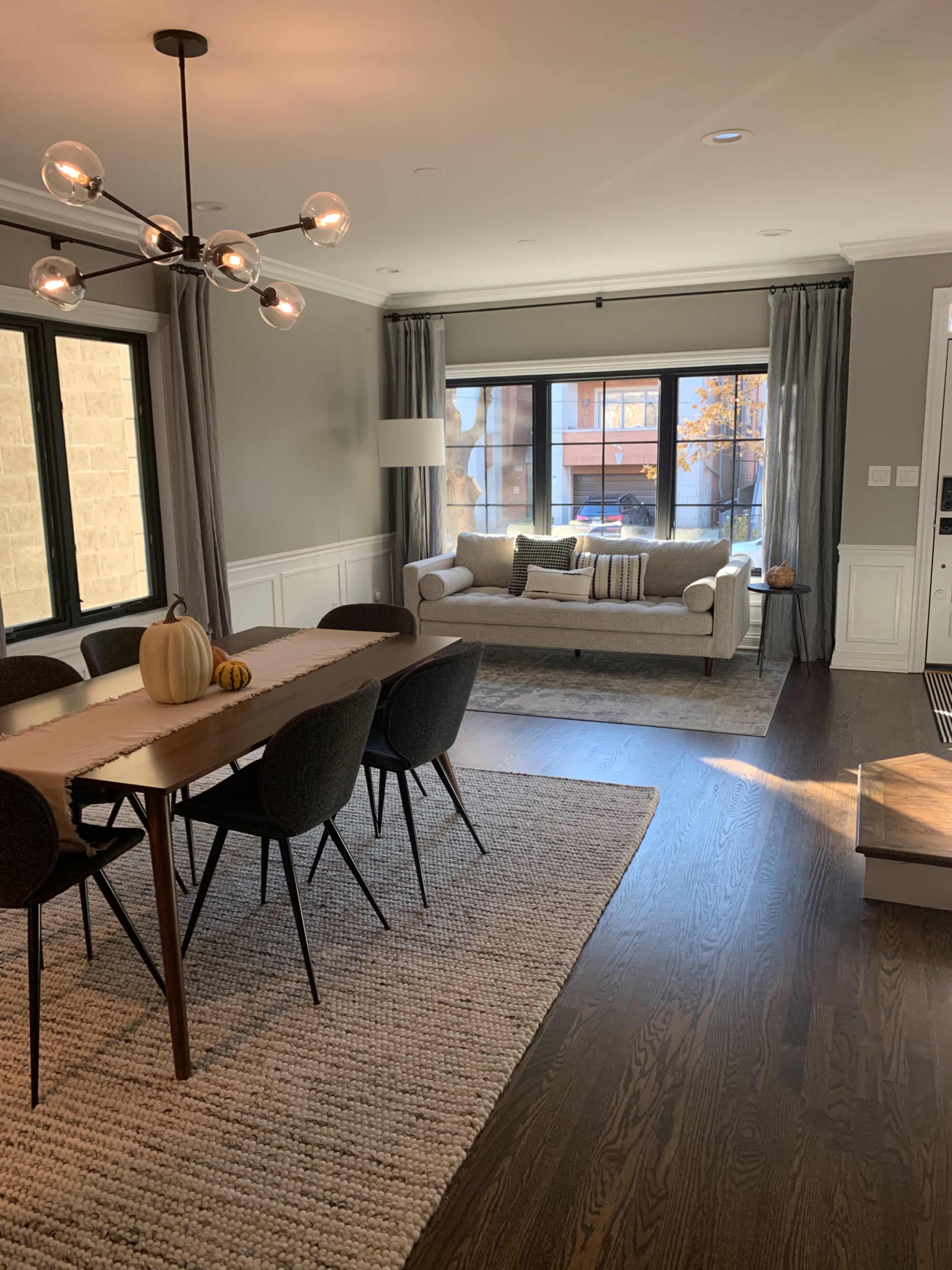 A dining area with a wooden table and chairs is adjacent to a living space featuring a sofa and large windows.
