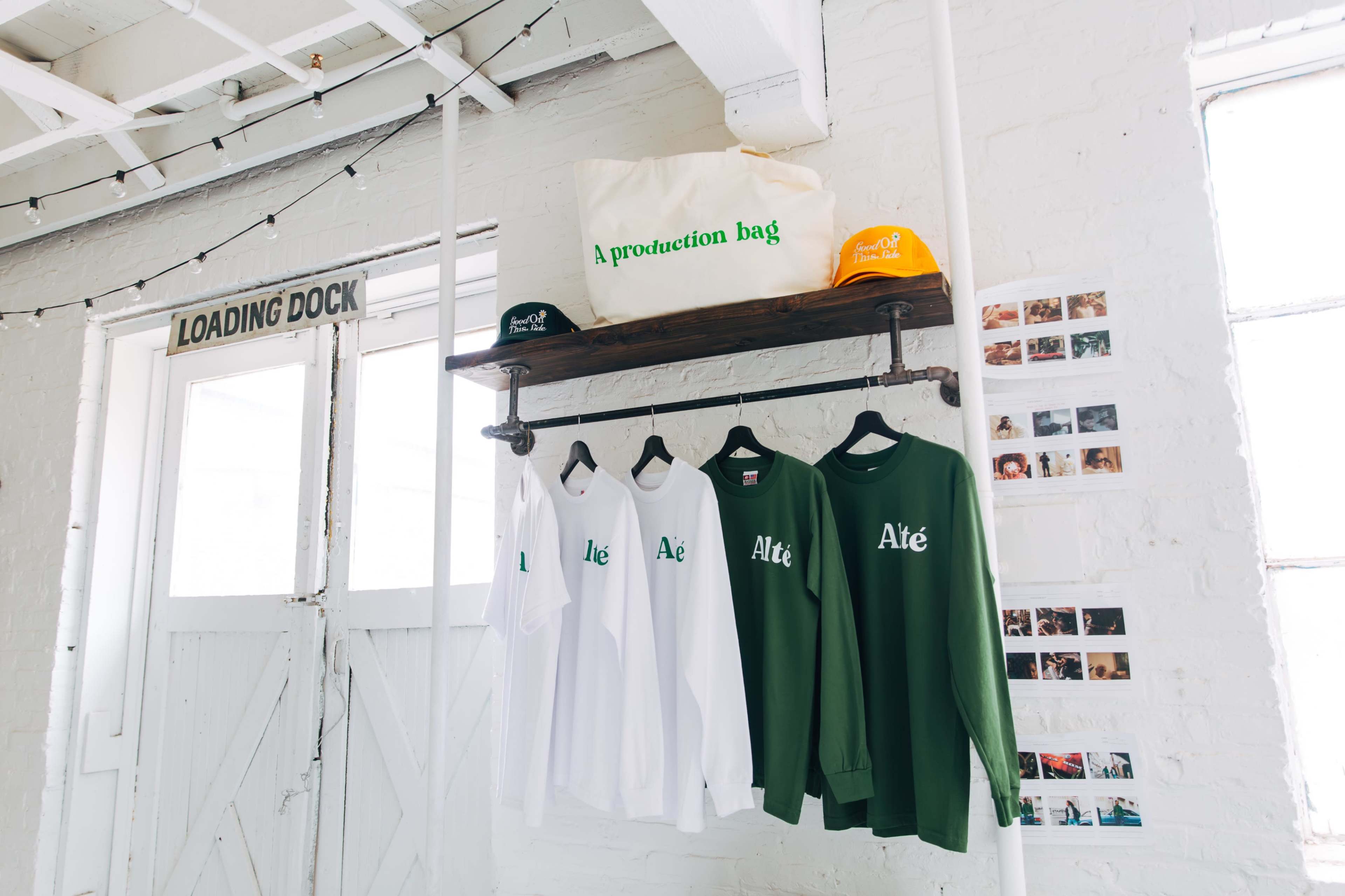 A display of clothing, including white and green shirts, hangs on a wooden rack next to a tote bag and hats in a bright, industrial space.