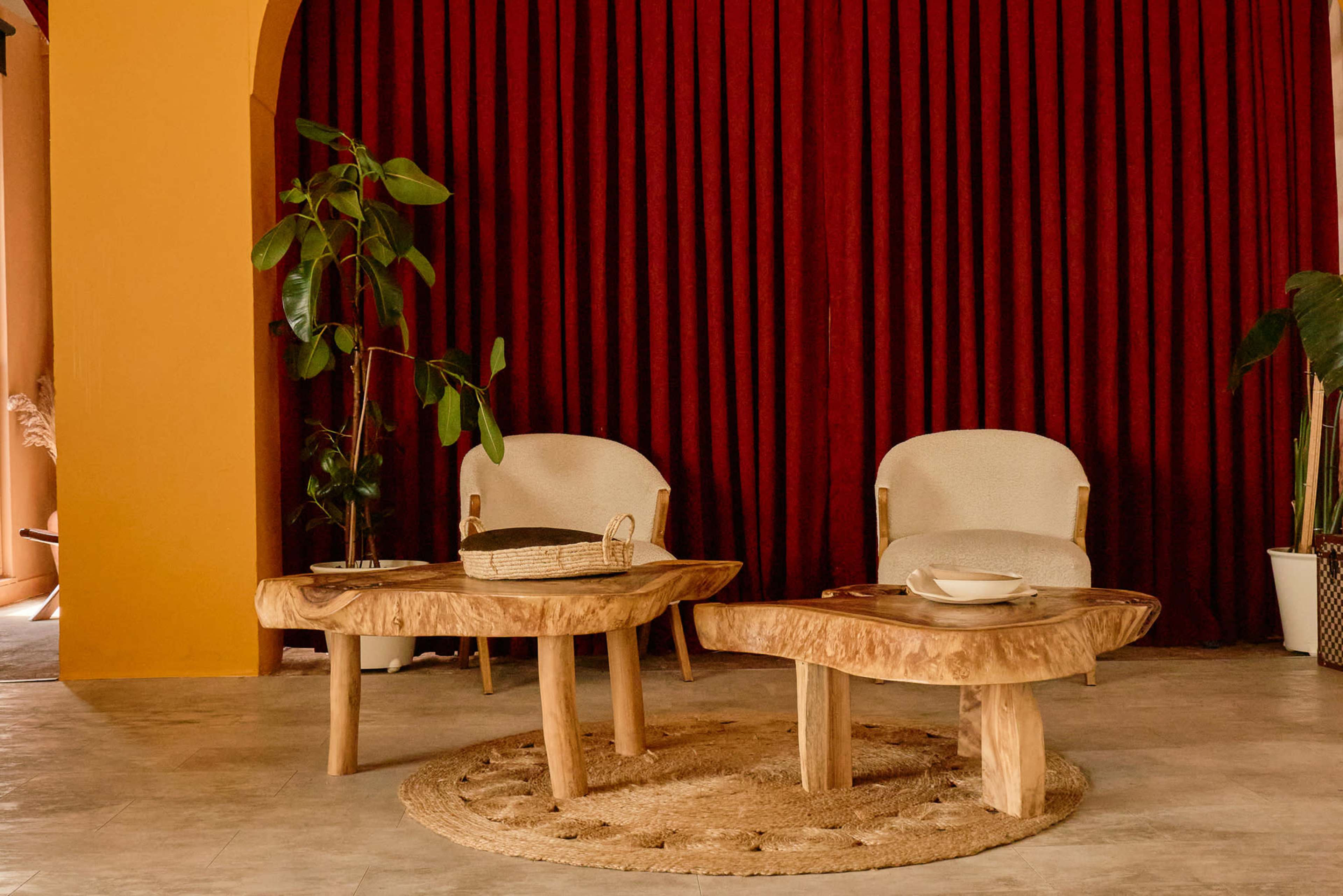The image depicts a cozy seating area with two upholstered chairs, a wooden coffee table, and a circular jute rug, set against a backdrop of rich red curtains.