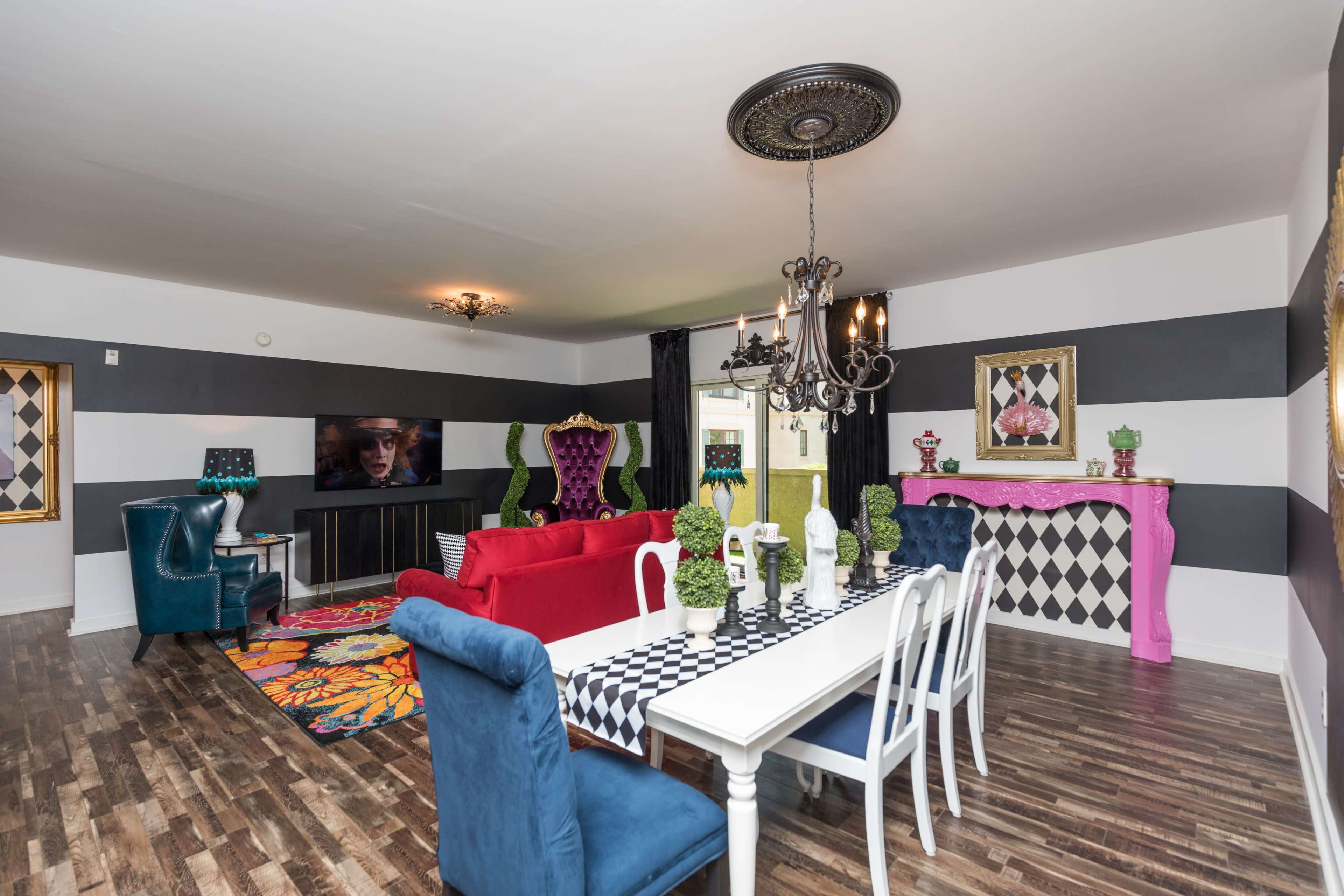 The living room features a mix of colorful furniture, including a red sofa and blue chairs, with striped walls, a large dining table, and various decorative elements.